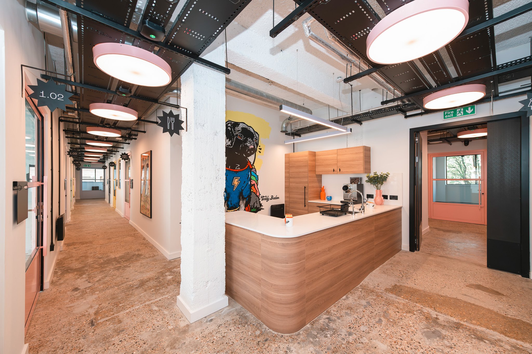 Modern office kitchen with wooden cabinets, white countertop, colorful artwork of a dog, and hallway with art portraits, black pendant lights, and pink doors.