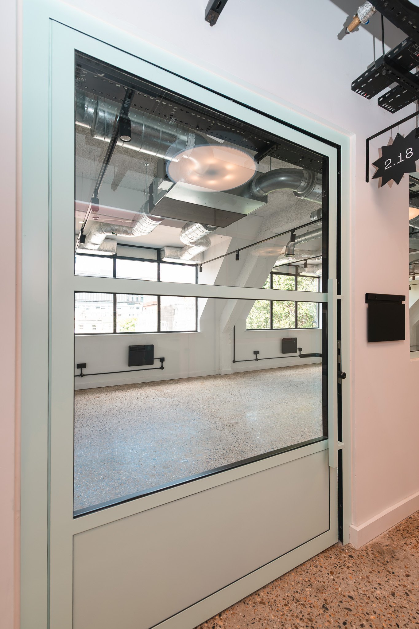 Modern interior office space with large windows, visible ceiling ductwork, and polished concrete floors, seen through a glass door.