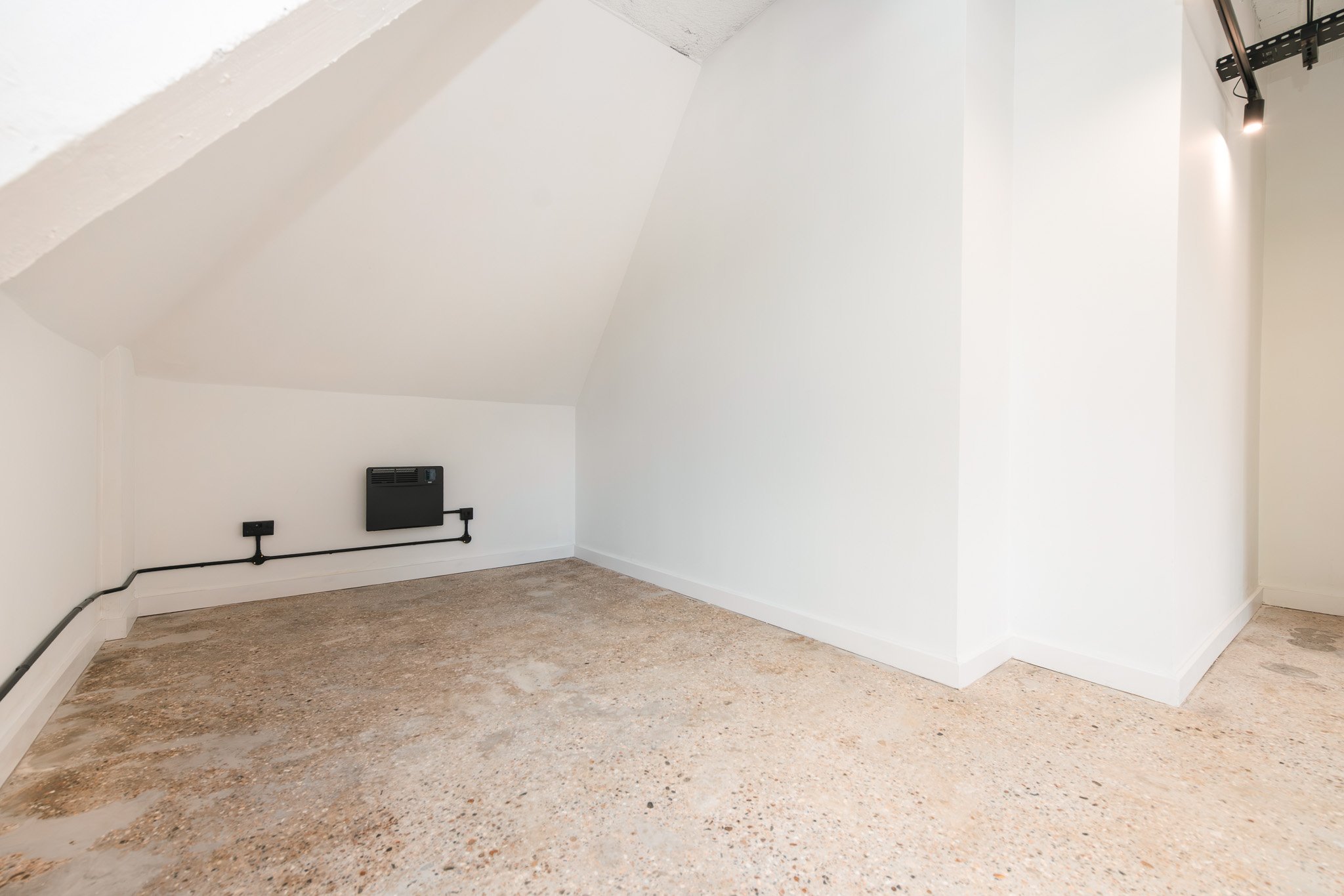 Empty room with sloped ceiling, white walls, exposed electrical wiring, and a heating unit on the wall.