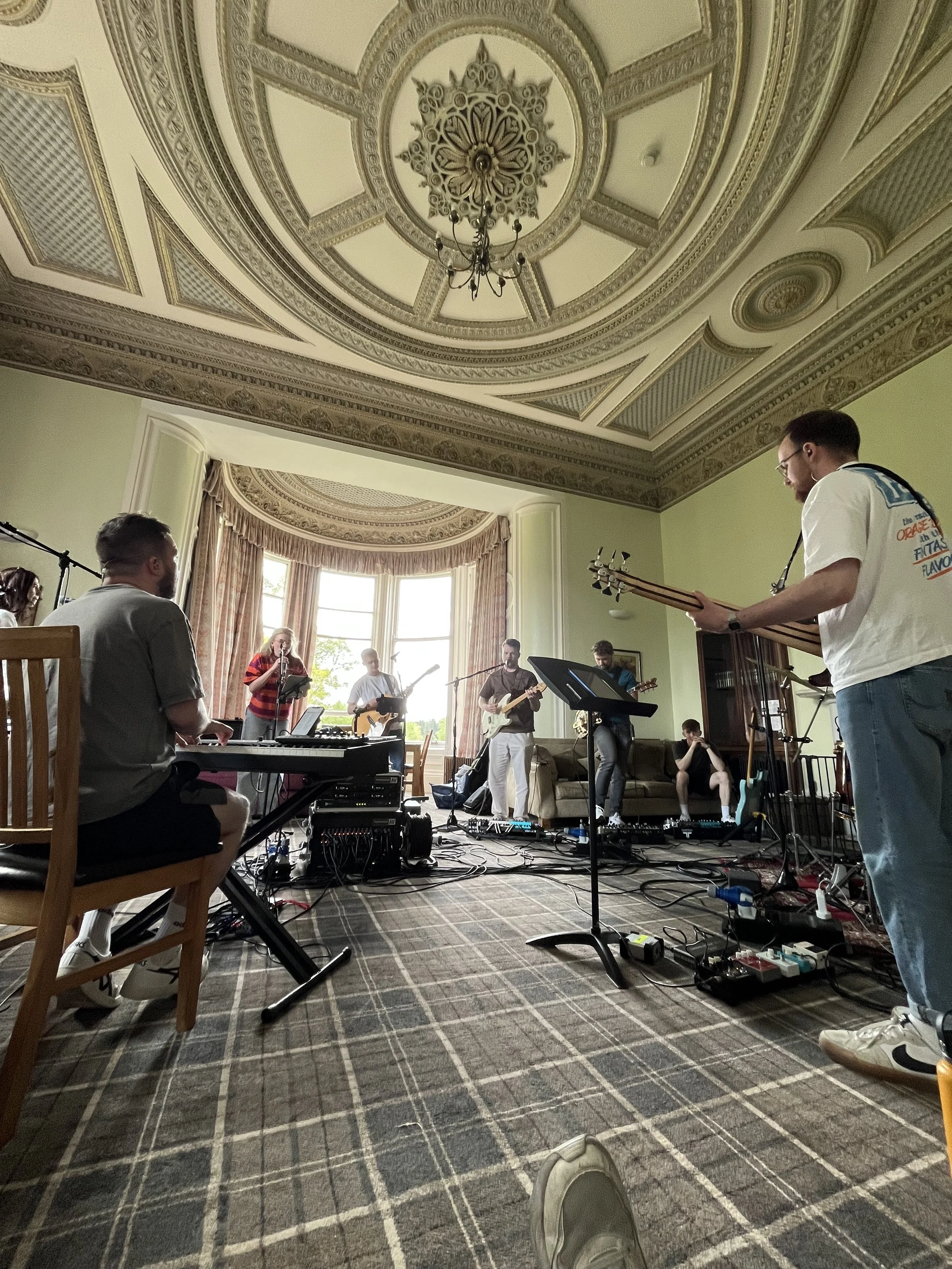 A group of musicians rehearsing in a large, ornate room with tall bay windows, high decorated ceilings, and a patterned carpet.