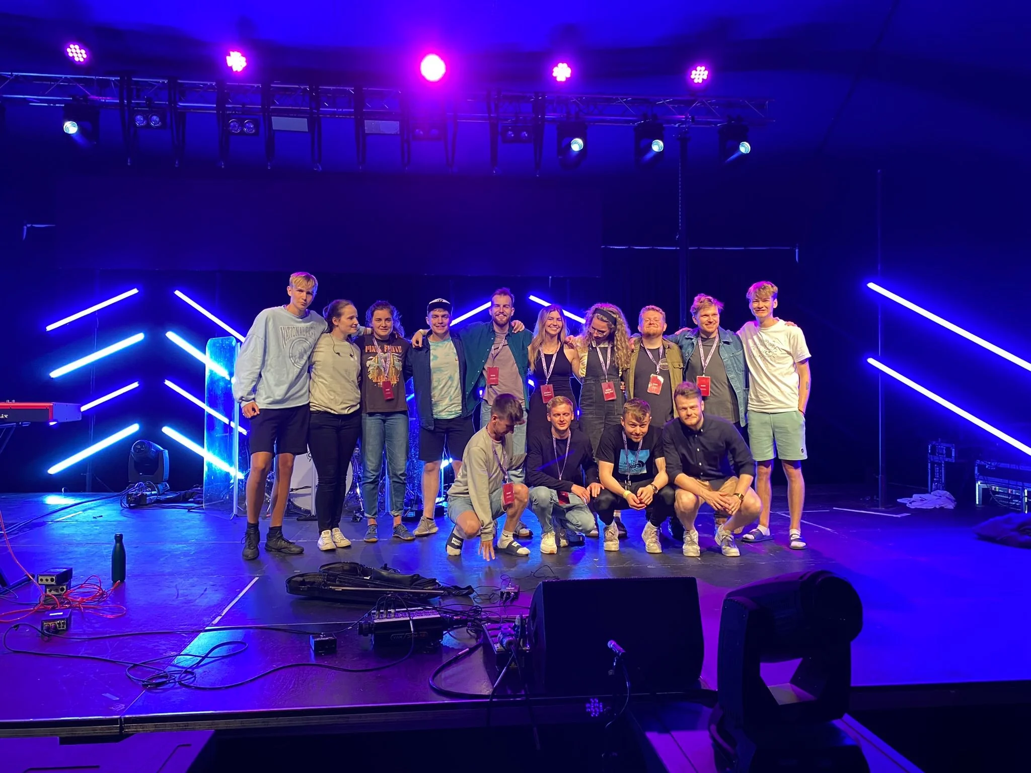 A group of people on stage at an event, posing for a photo with neon lights in the background. Some are kneeling in the front row, while others are standing behind.