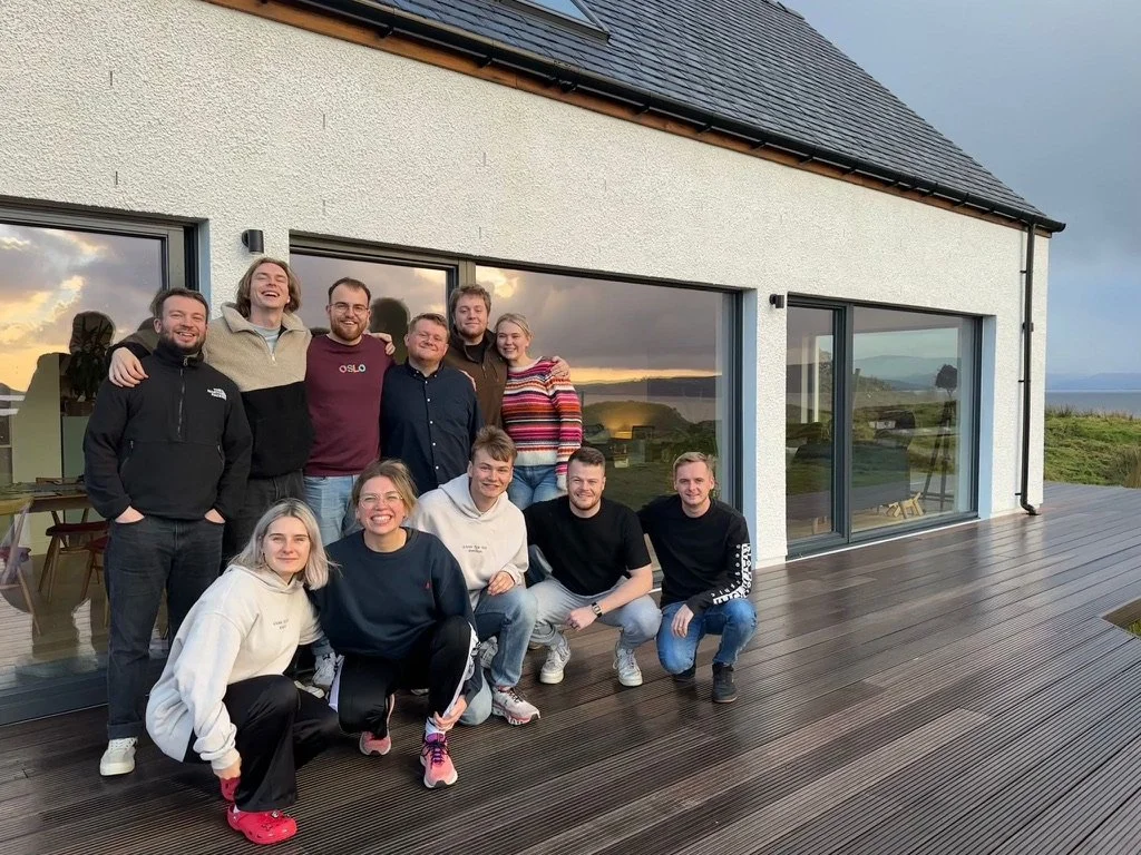 Group of eleven people smiling and posing in front of a house with large windows, wooden deck, and a scenic landscape background during sunset.