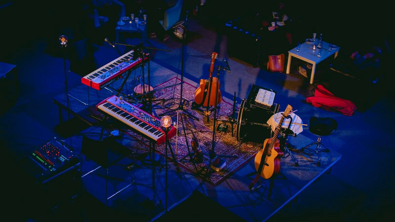 Stage set with musical instruments including two keyboards, an acoustic guitar, a small drum machine, an amplifier, and microphones, with a red patterned rug and audience seated in the background.