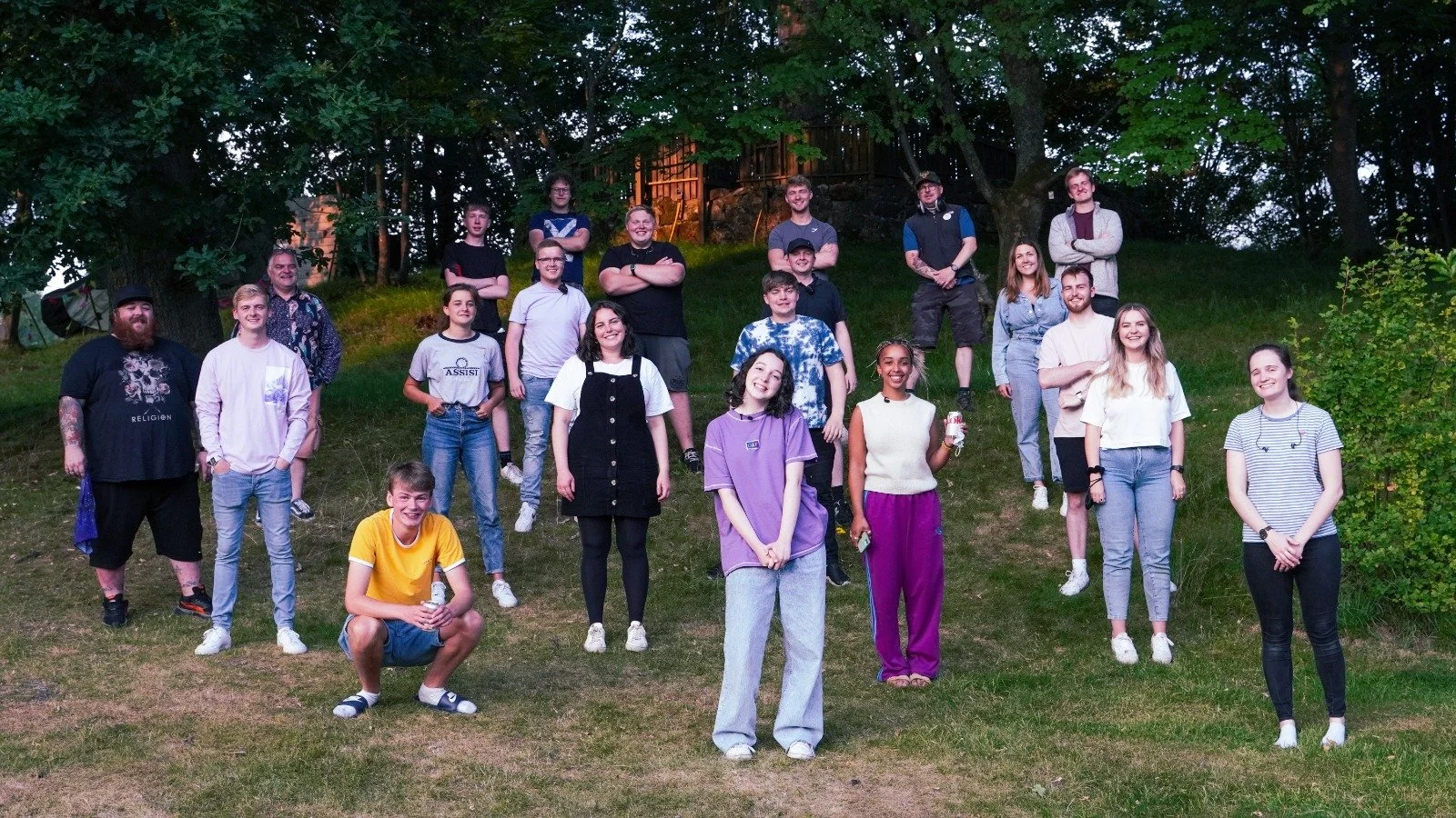 A group of 18 young people and one older person posing outdoors on a grassy hillside, with trees and a wooden structure in the background during dusk.