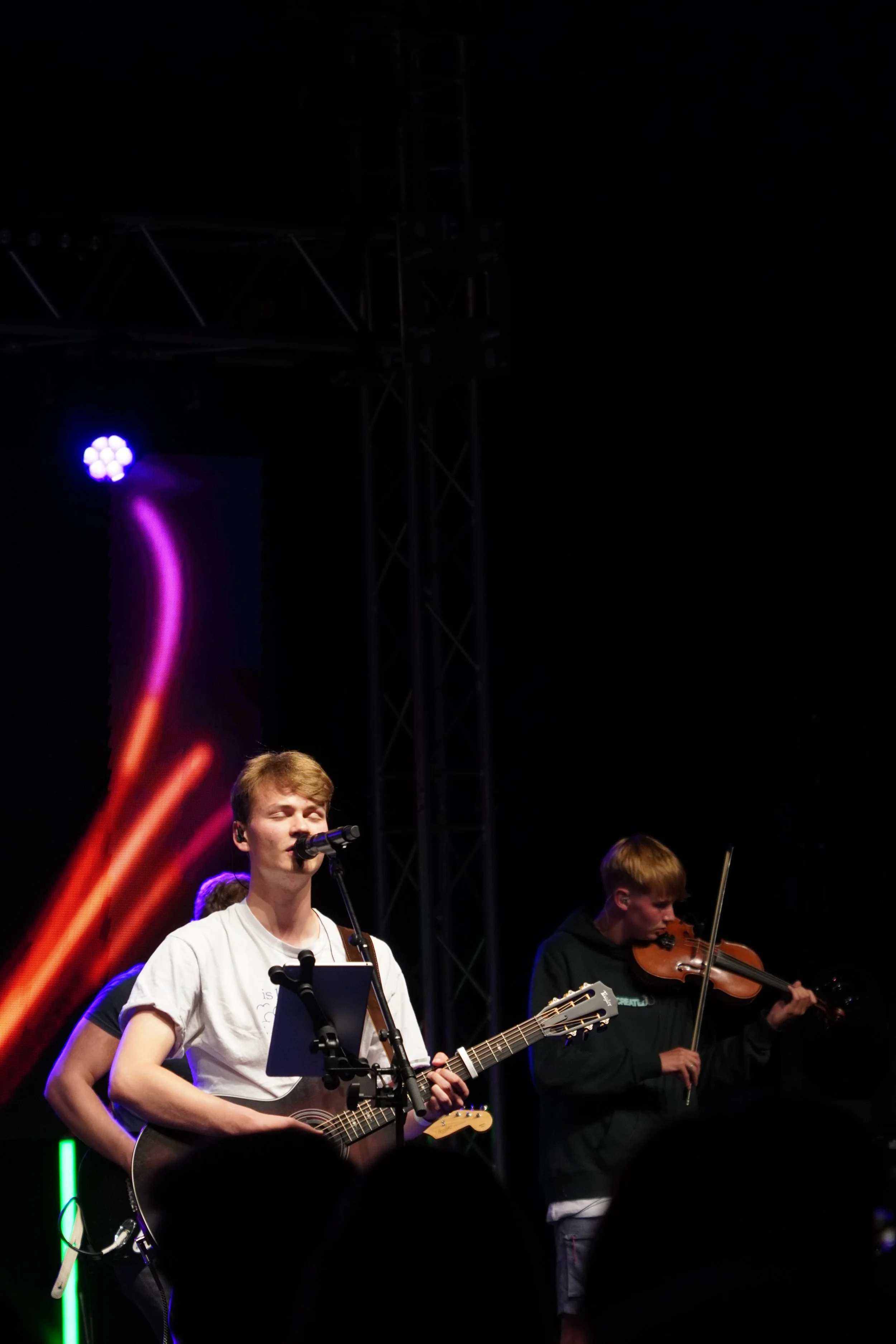 Young male singer playing an acoustic guitar and singing into a microphone, with a young male violinist beside him, performing on stage at night with colorful stage lights.