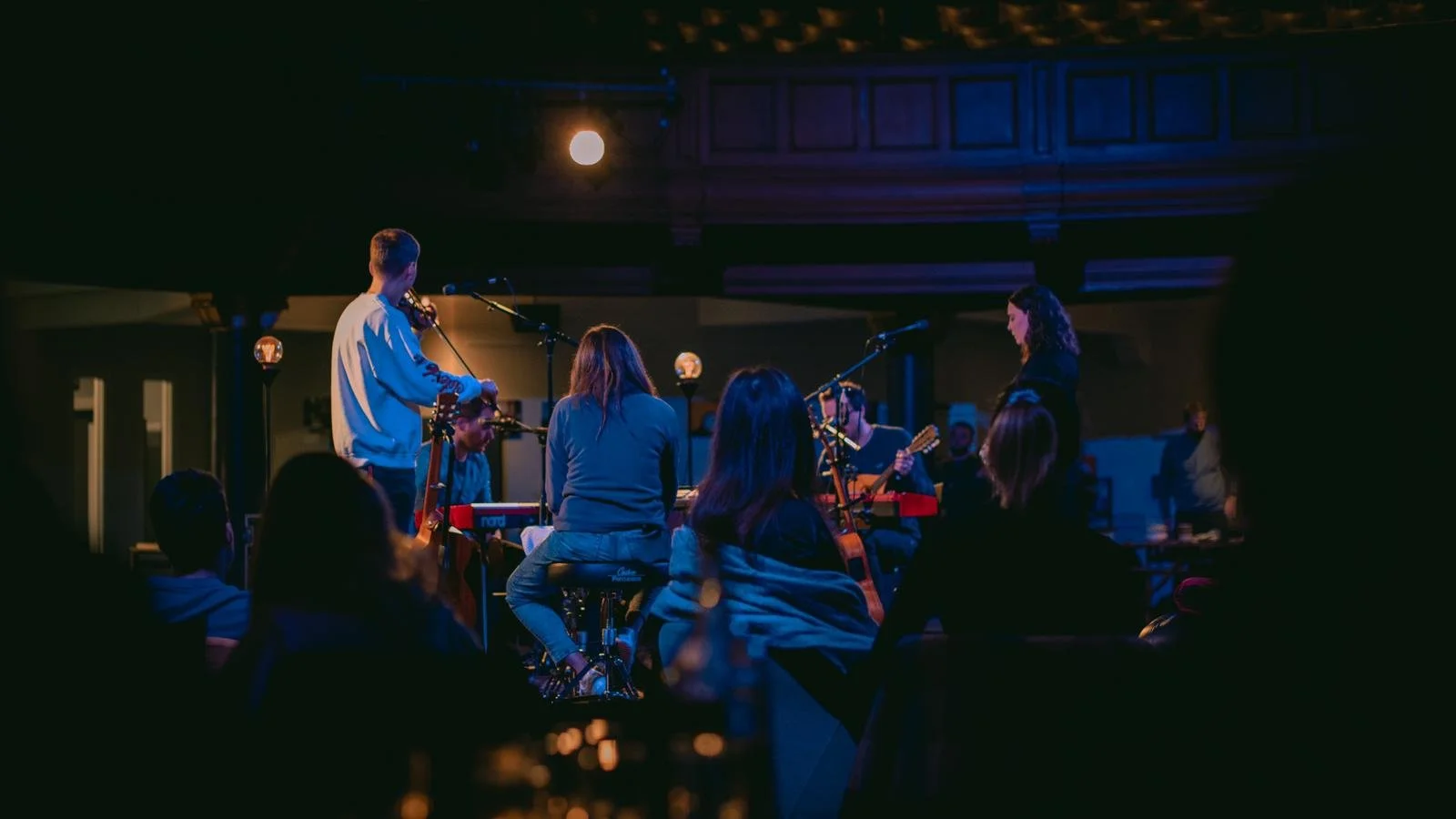 A live musical performance featuring a band with guitarists, a keyboardist, a vocalist, and an audience in a dimly lit venue with blue and purple lighting.