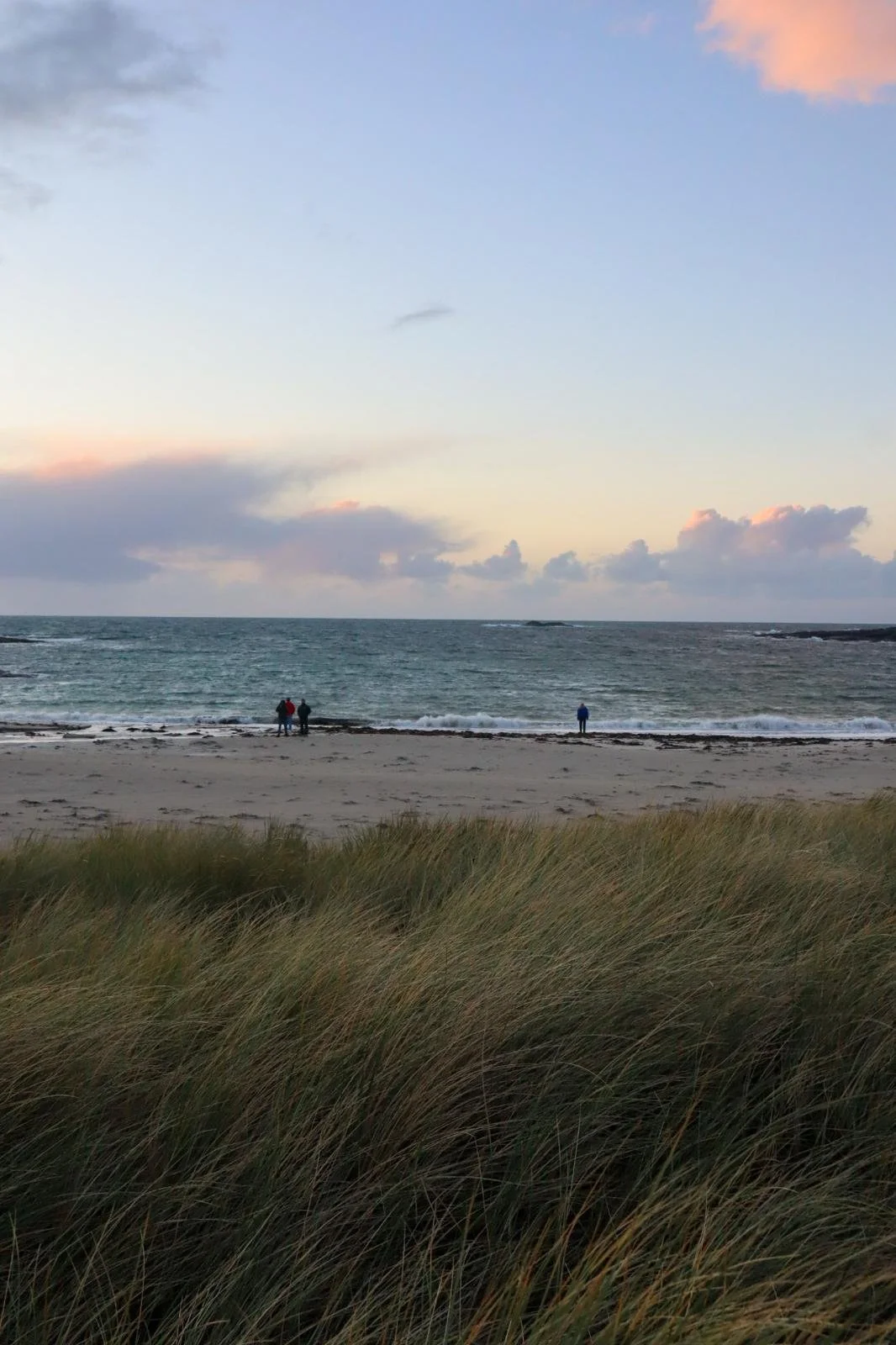 Beach scene at sunset with sand dunes in the foreground, three people standing near the water, and a partly cloudy sky with pink and orange hues.