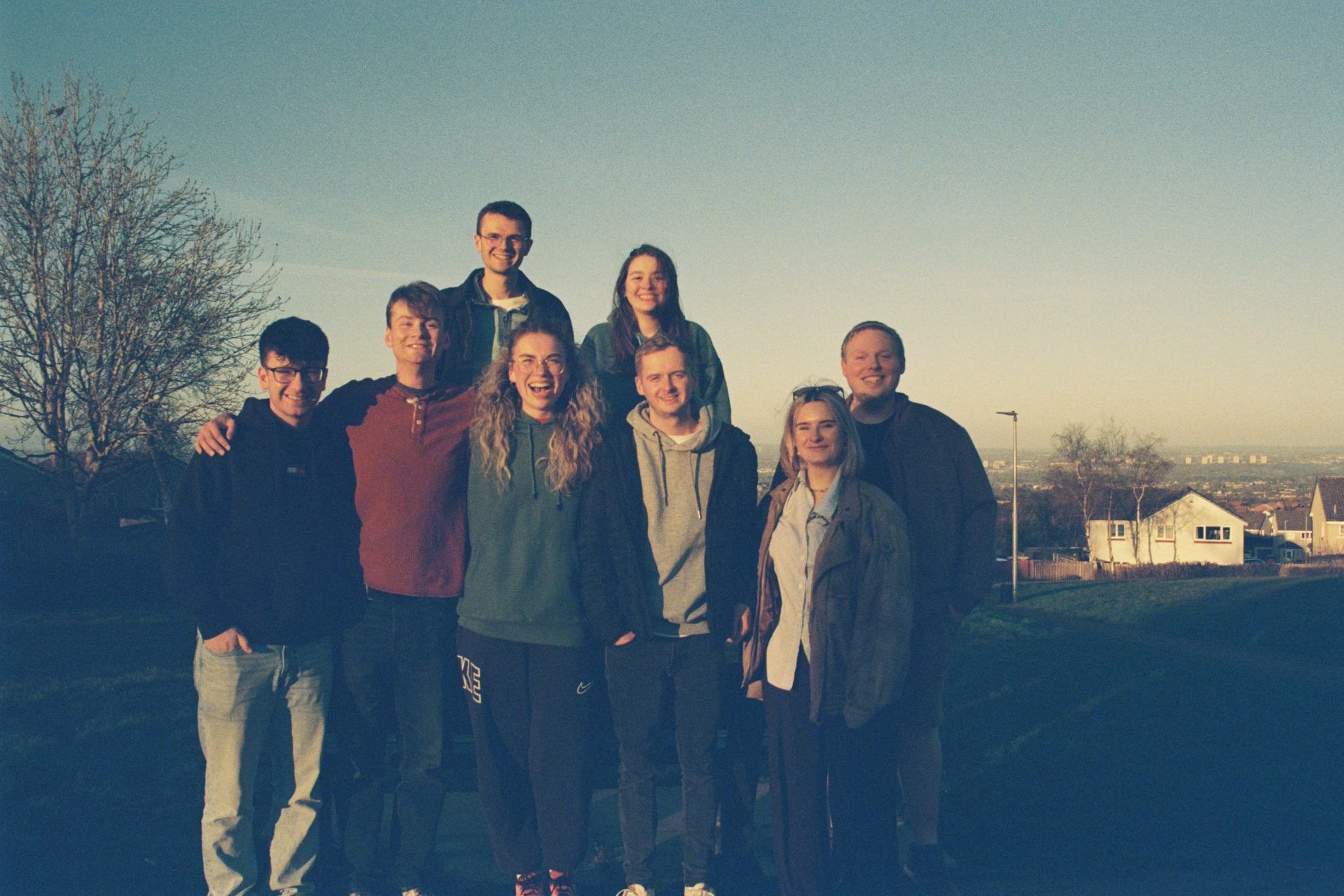 Group of nine young people standing outdoors during sunset, smiling with houses and trees in the background.