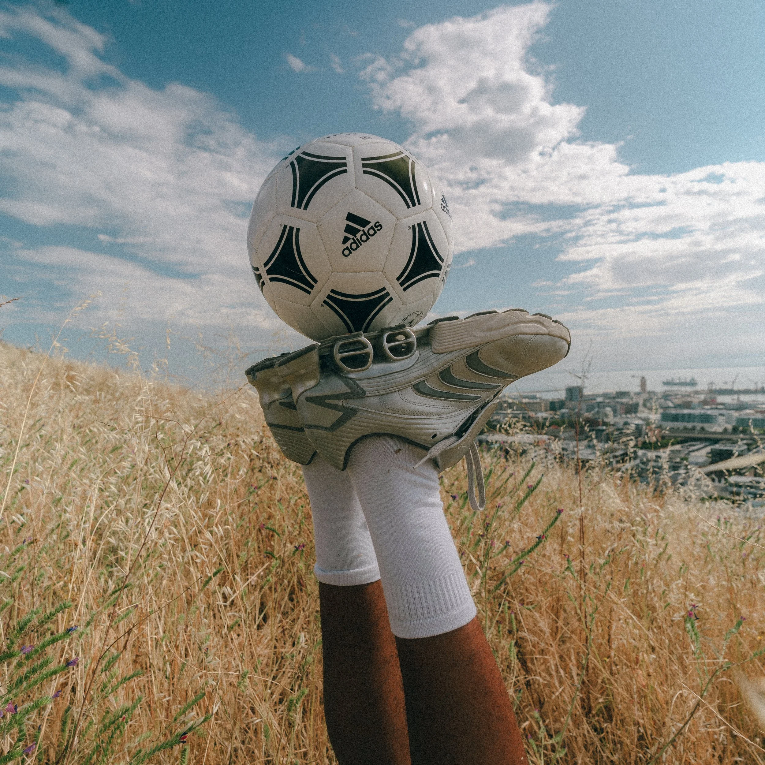 A person wearing a white sock and a gray sneaker balancing a soccer ball on their finger in a grassy field with a cityscape in the background and clouds in the sky.