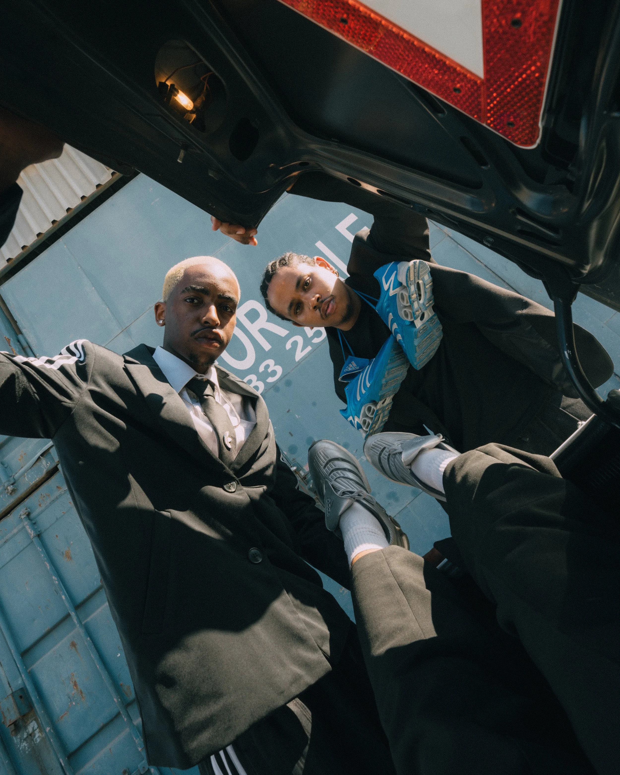 Two men and a woman looking down into the camera from inside a vehicle, with a sign in the background reading 'FUR 33 25'. They are dressed in black suits, with one man wearing a black tie and the woman wearing Adidas football cleats, white socks, an