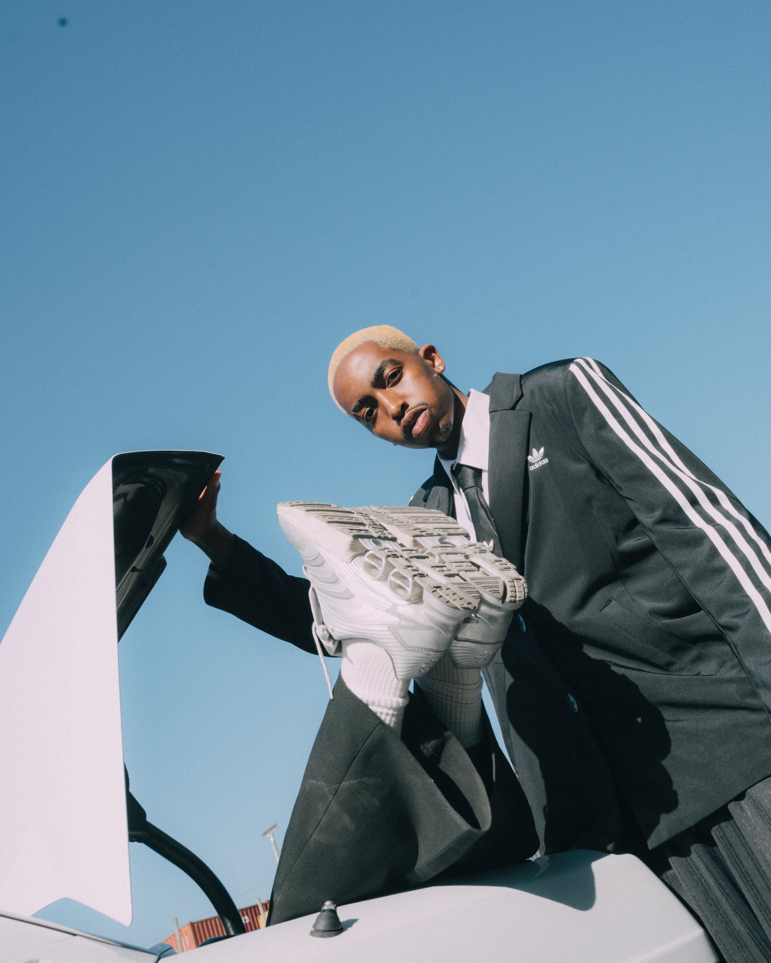 Man with platinum blonde hair wearing a black Adidas jacket with white stripes, holding a white sneaker, leaning over a white car with the sunroof open against a clear blue sky.