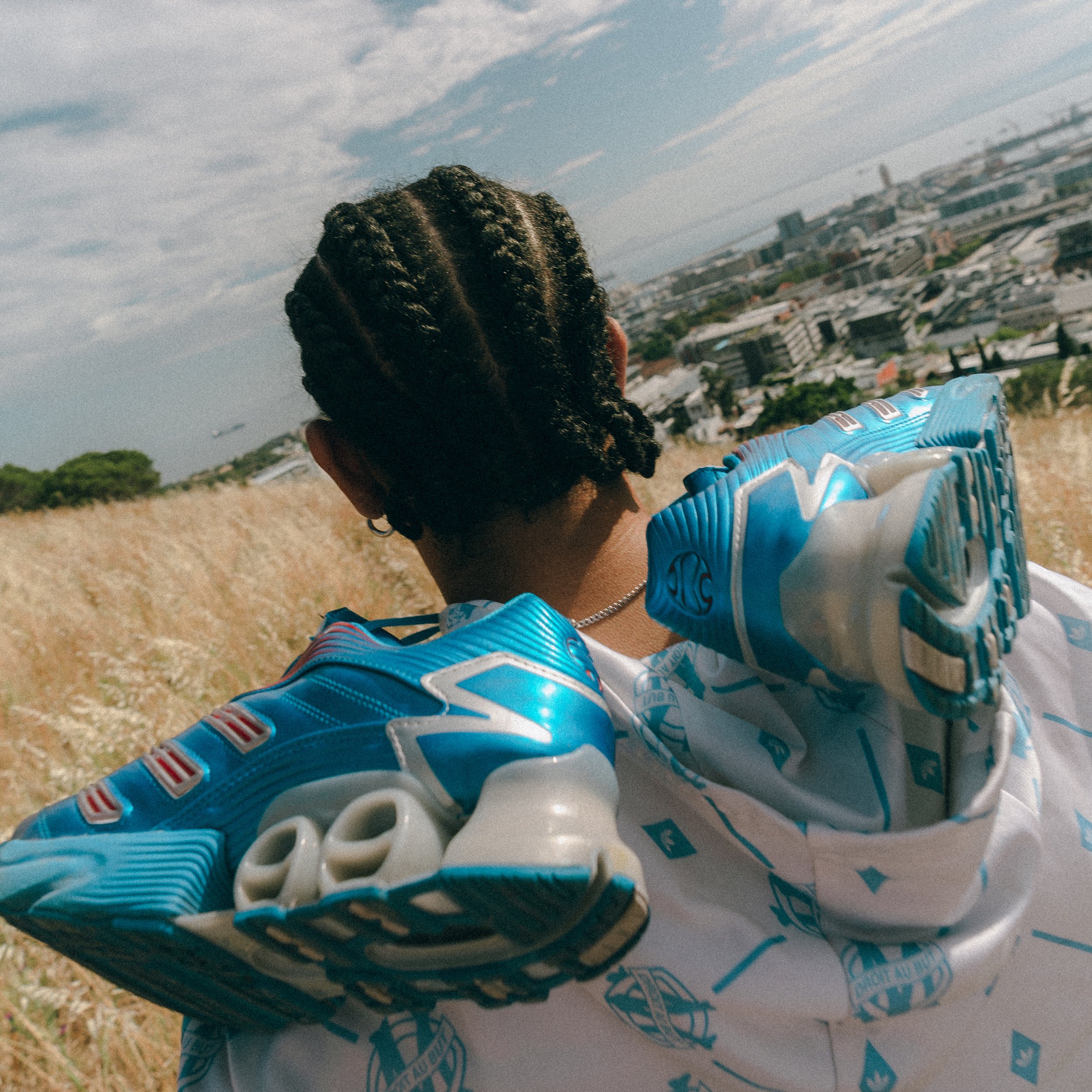 Person wearing blue and white sneakers and white hoodie with blue patterns, standing on a hill overlooking a cityscape and water in the distance.