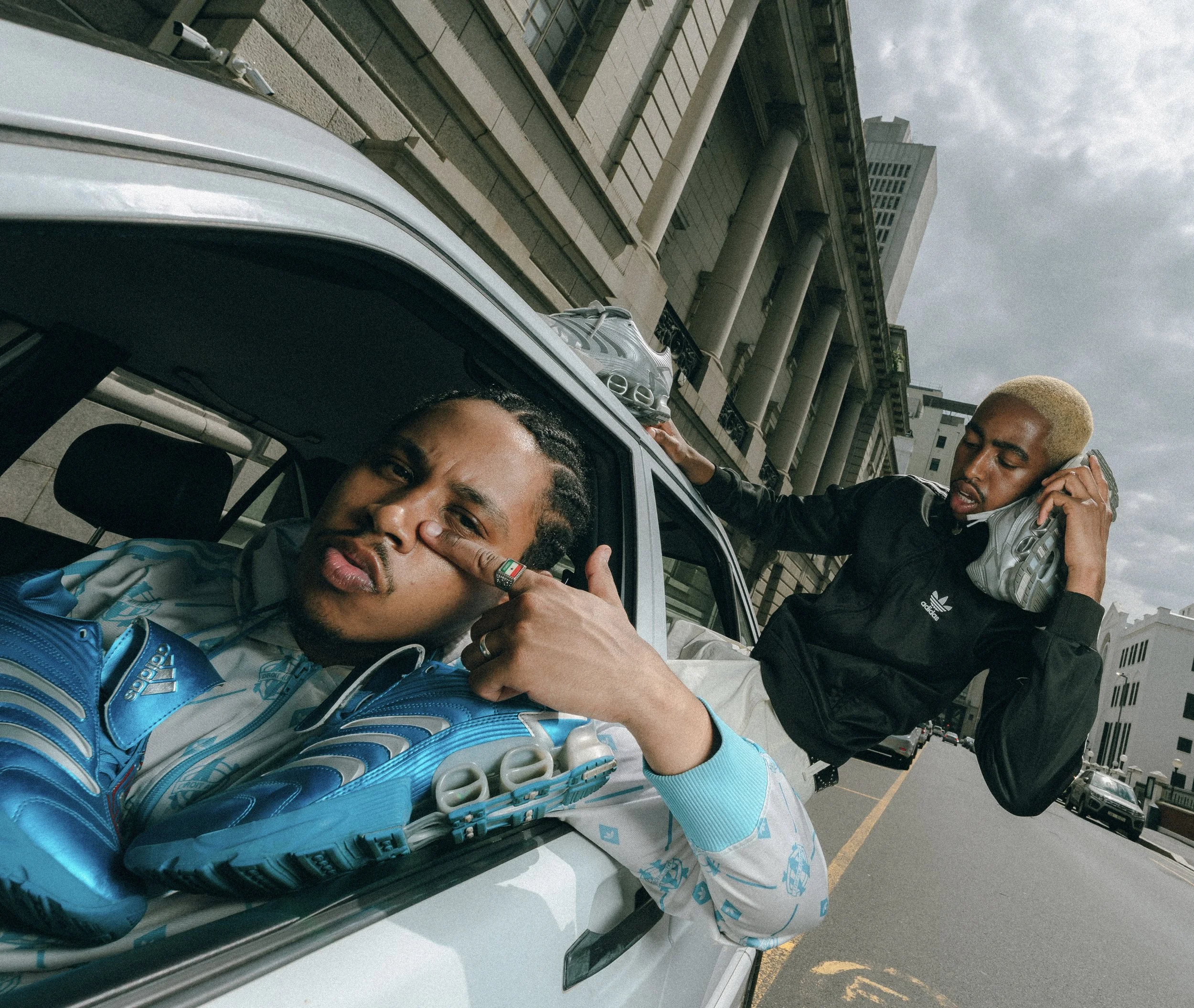 Two young men with trendy hairstyles and casual sportswear, one inside a car and the other on the street, posing with a serious and confident attitude amid an urban cityscape.