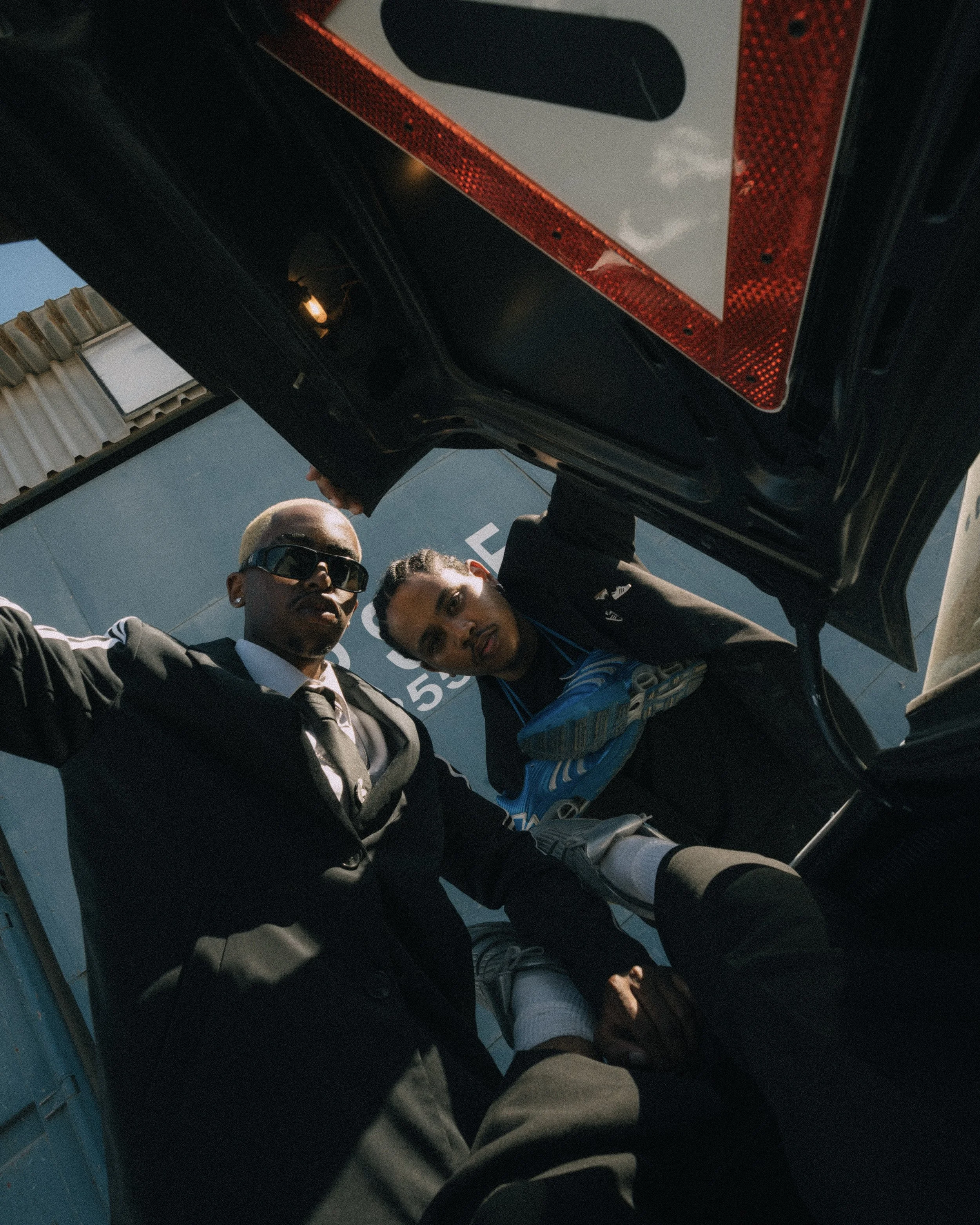 Two young men, one in a suit and tie with sunglasses and the other in athletic wear with a sneaker, look down into the camera from an open trunk of a car during daytime.