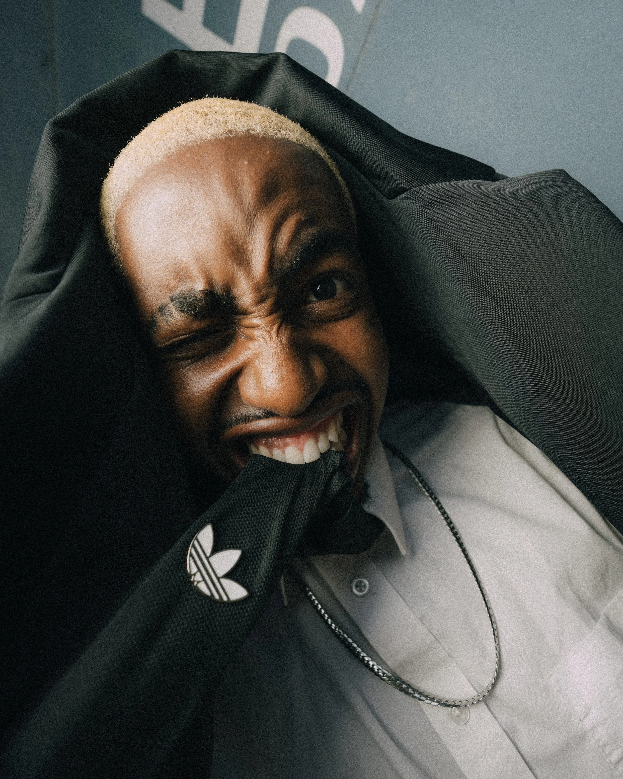 A man with light blonde hair making a funny face, holding his head with one hand, wearing a white shirt, silver chain, and black Adidas jacket.