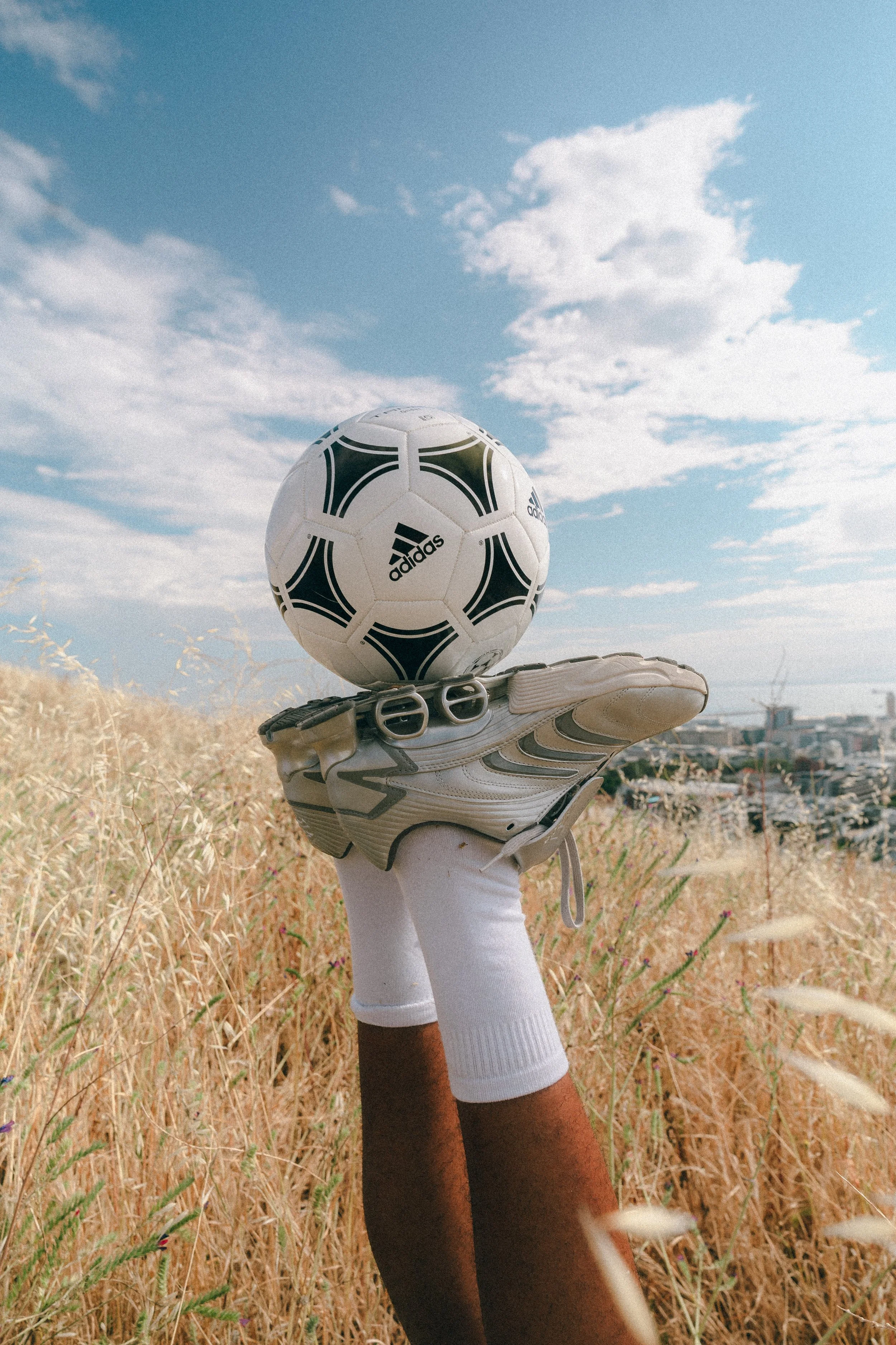 A hand wearing a white glove holding a soccer ball on a grassy hill with a cityscape in the background under a blue sky with clouds.