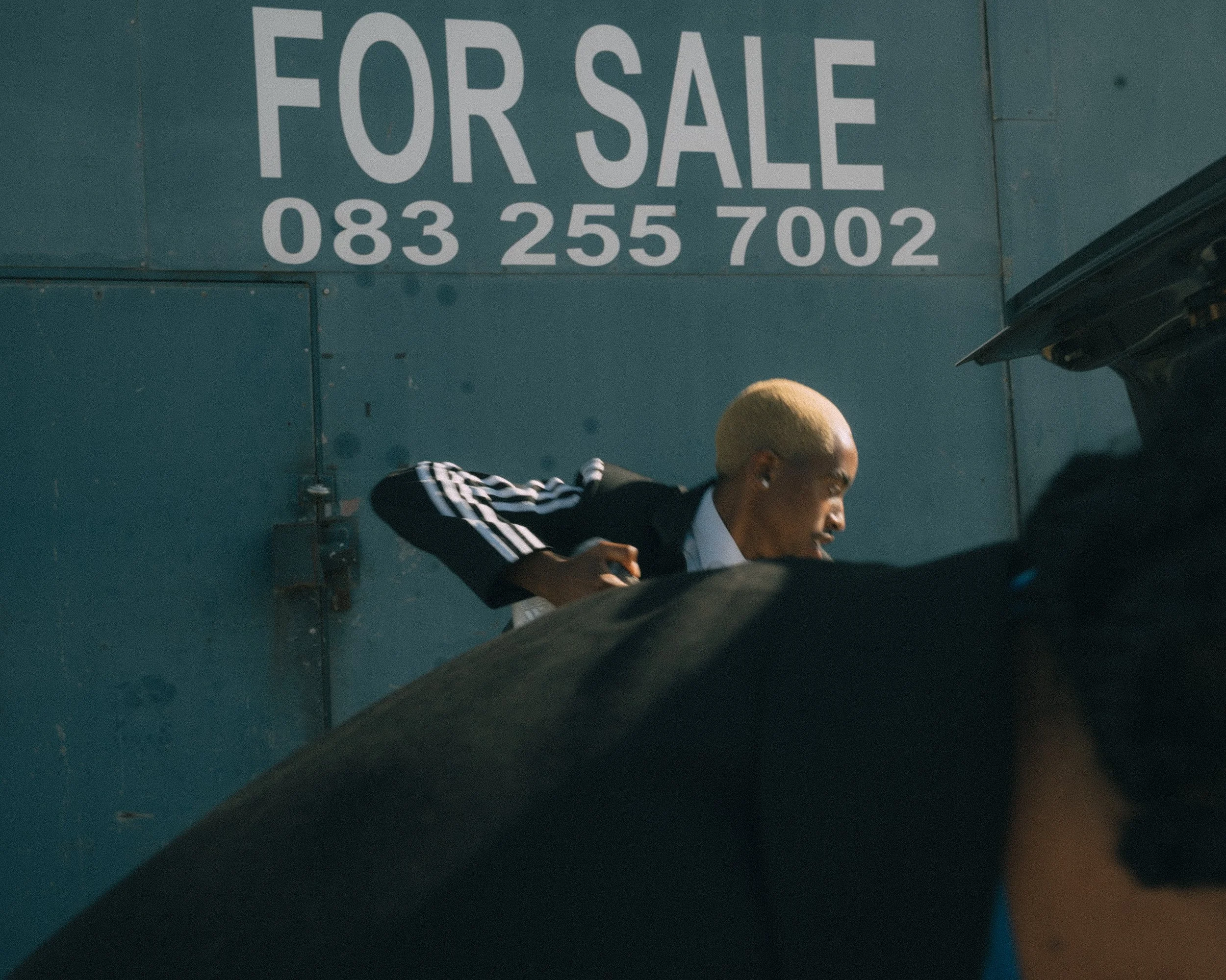 A person with bleached blonde hair wearing a black and white Adidas jacket bending over a car engine, with a blue wall and a 'For Sale' sign and phone number in the background.