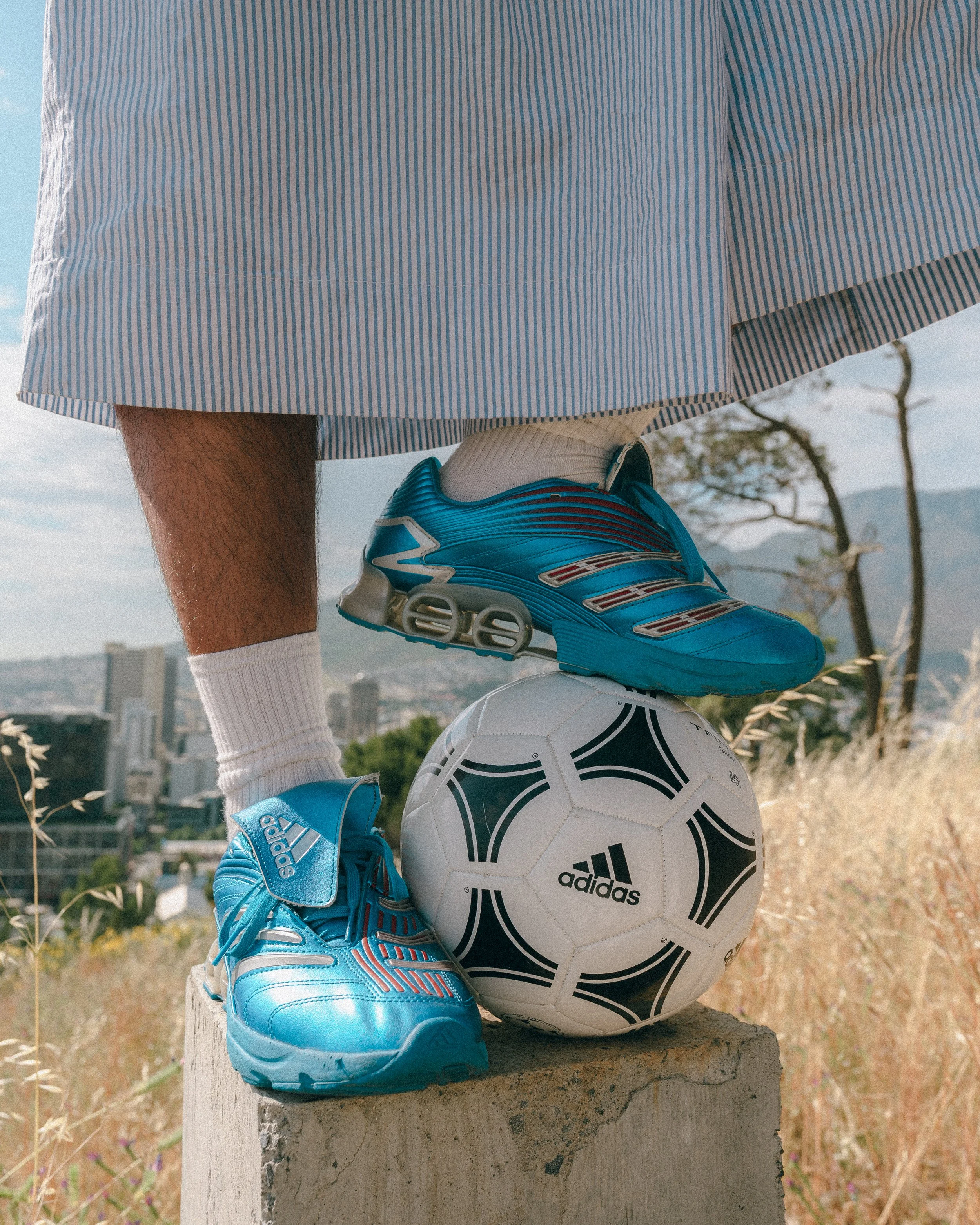 A person balancing on a concrete block with one foot, wearing blue running shoes, a white sock, and a striped blue and white skirt. An Adidas soccer ball is underneath the foot.