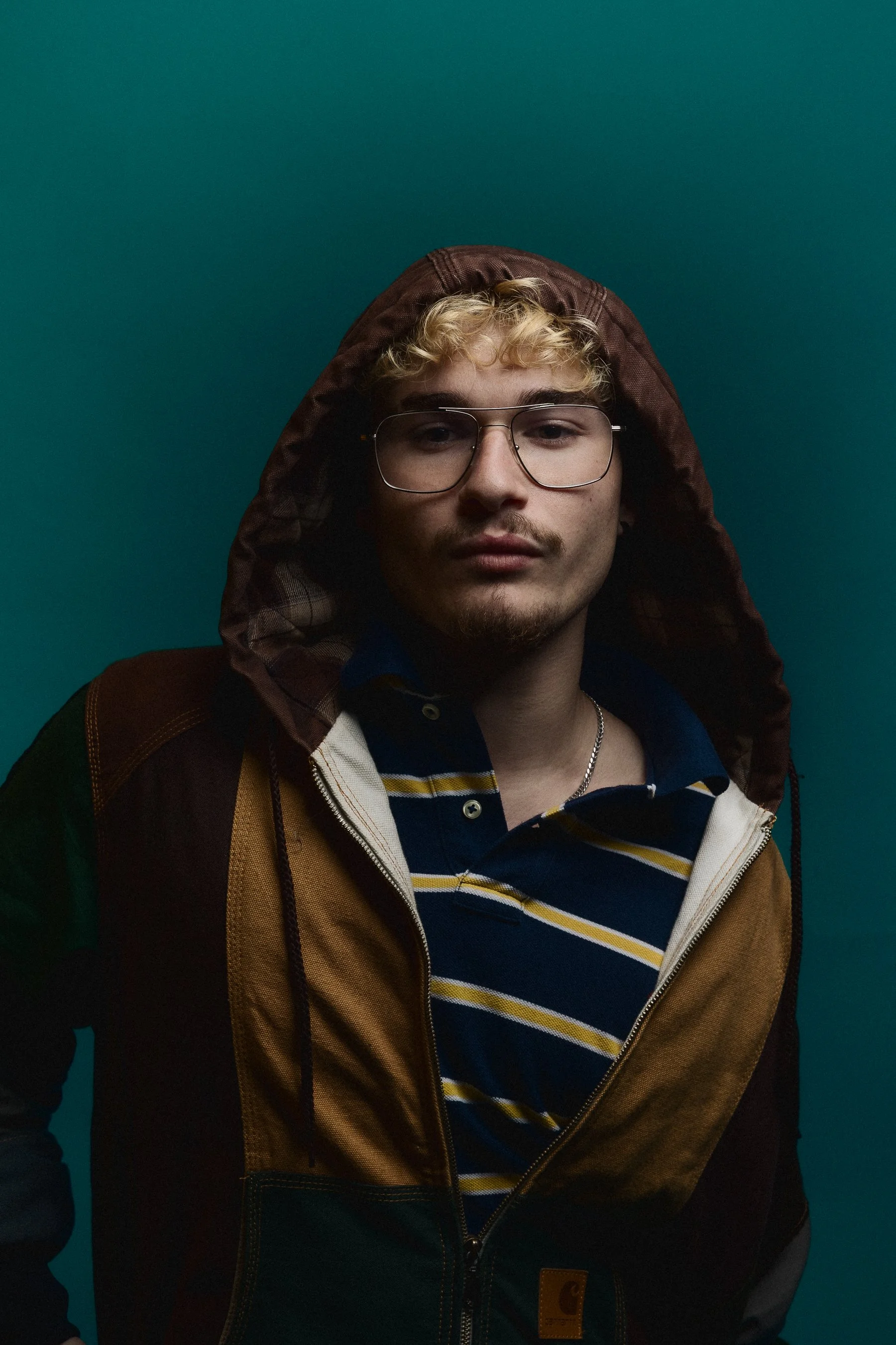 Close-up portrait of a young man wearing glasses and hooded jacket against a teal background.