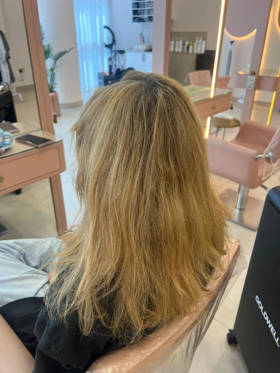 A woman with blonde, shoulder-length hair sitting in a salon chair inside a modern hair salon with pink and beige decor.