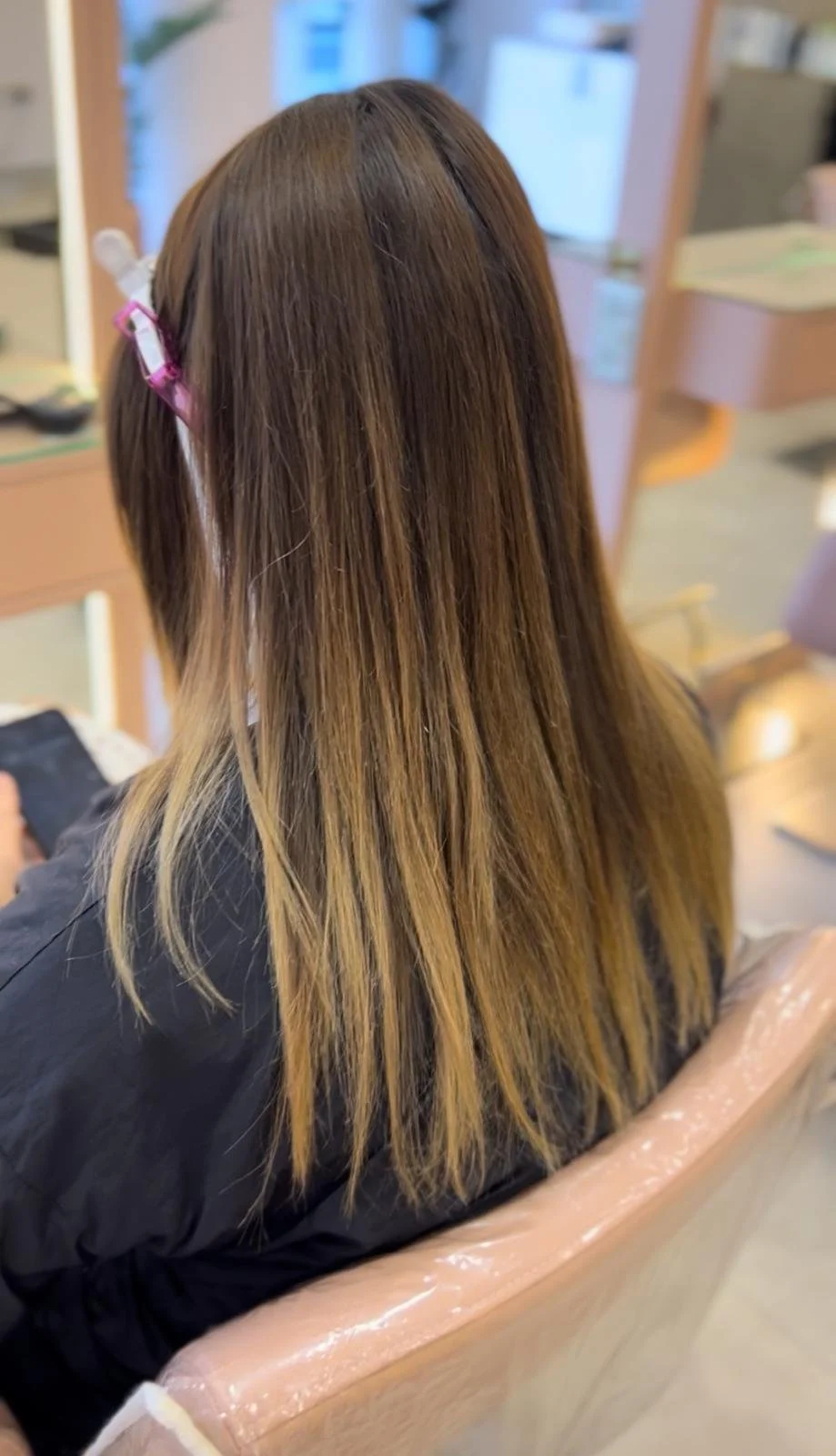Back view of a woman with long, straight, brown hair sitting in a salon chair with a cape around her shoulders.