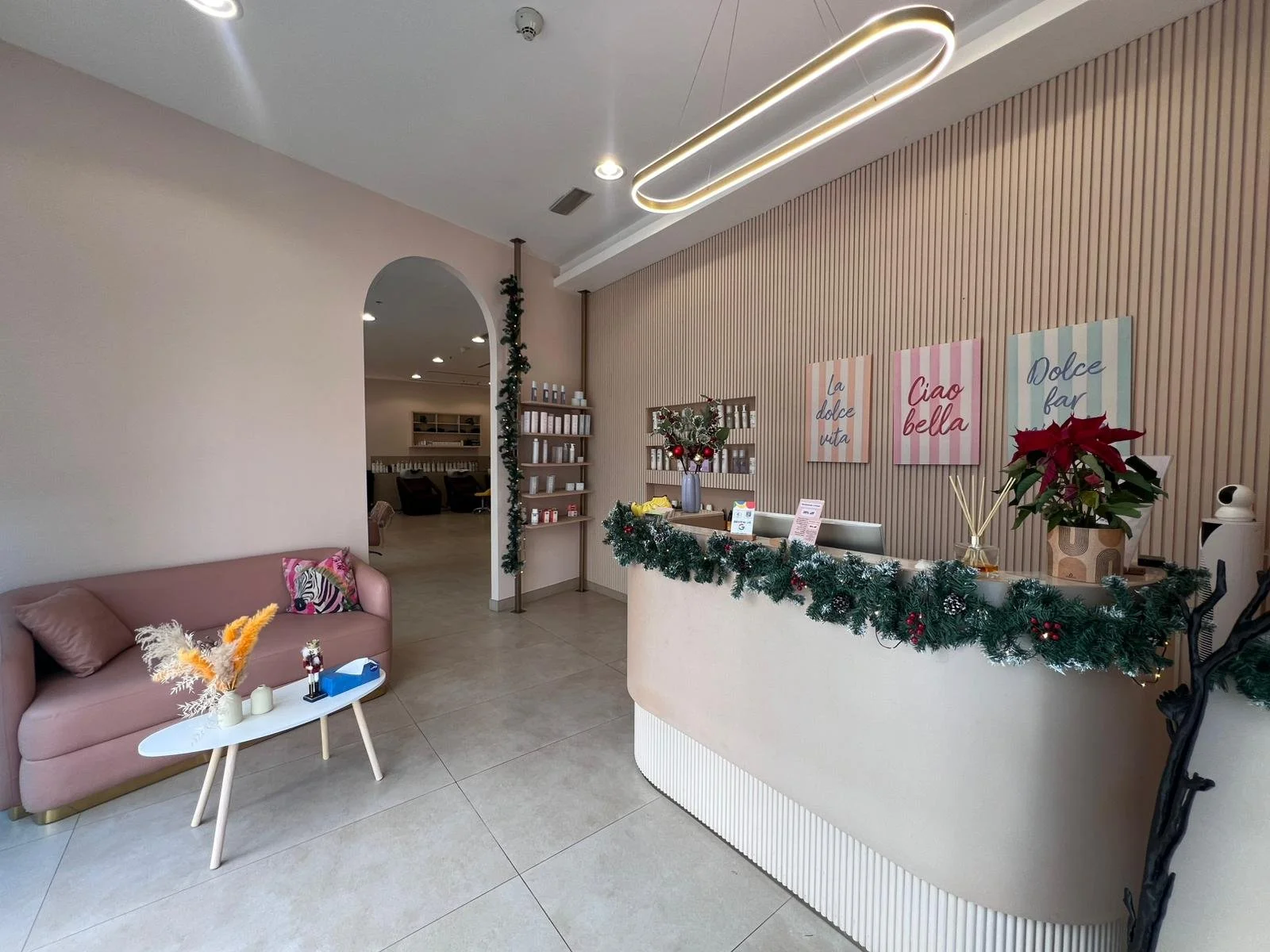 Decorated reception desk with Christmas garland, poinsettia, and holiday signs, pink sofa with pillow, small white coffee table with dried flowers, and a hallway with shelves, in a modern interior.