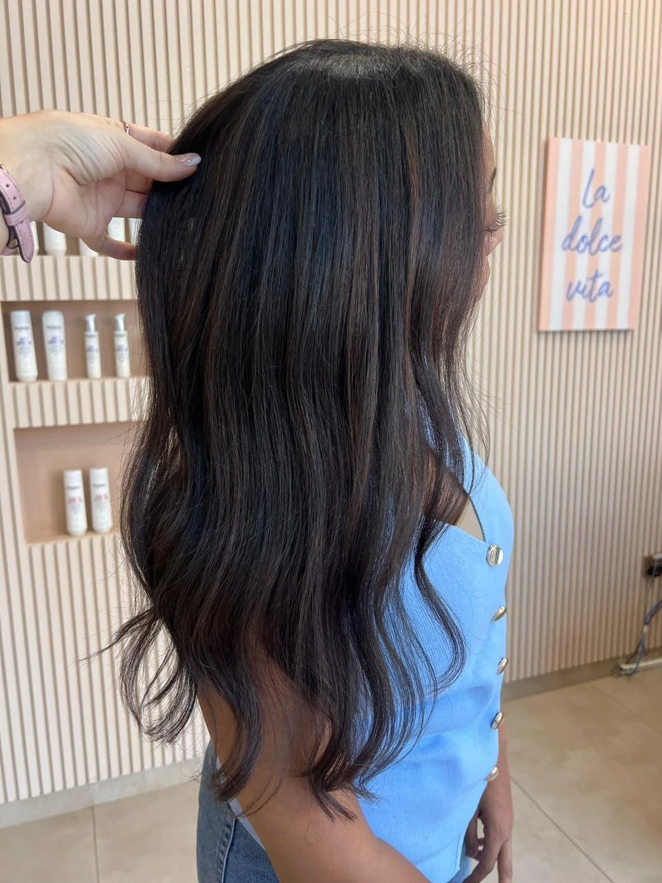 Side view of a woman with long, wavy brunette hair wearing a light blue top with gold buttons, in a salon with cream-colored walls and shelves with hair products.