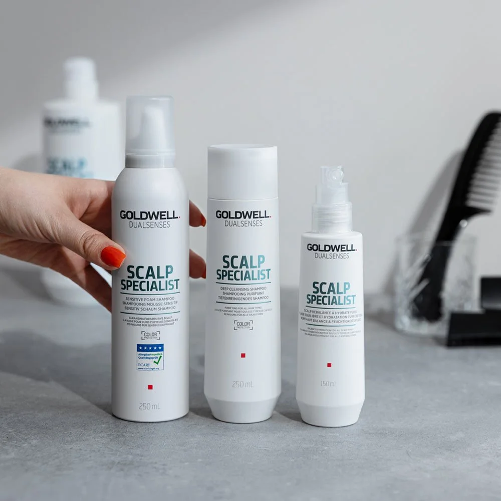 Three bottles of Goldwell Dualsenses Scalp Specialist shampoo on a gray surface, with a hand with red nail polish holding the left bottle, and a black comb in the background.
