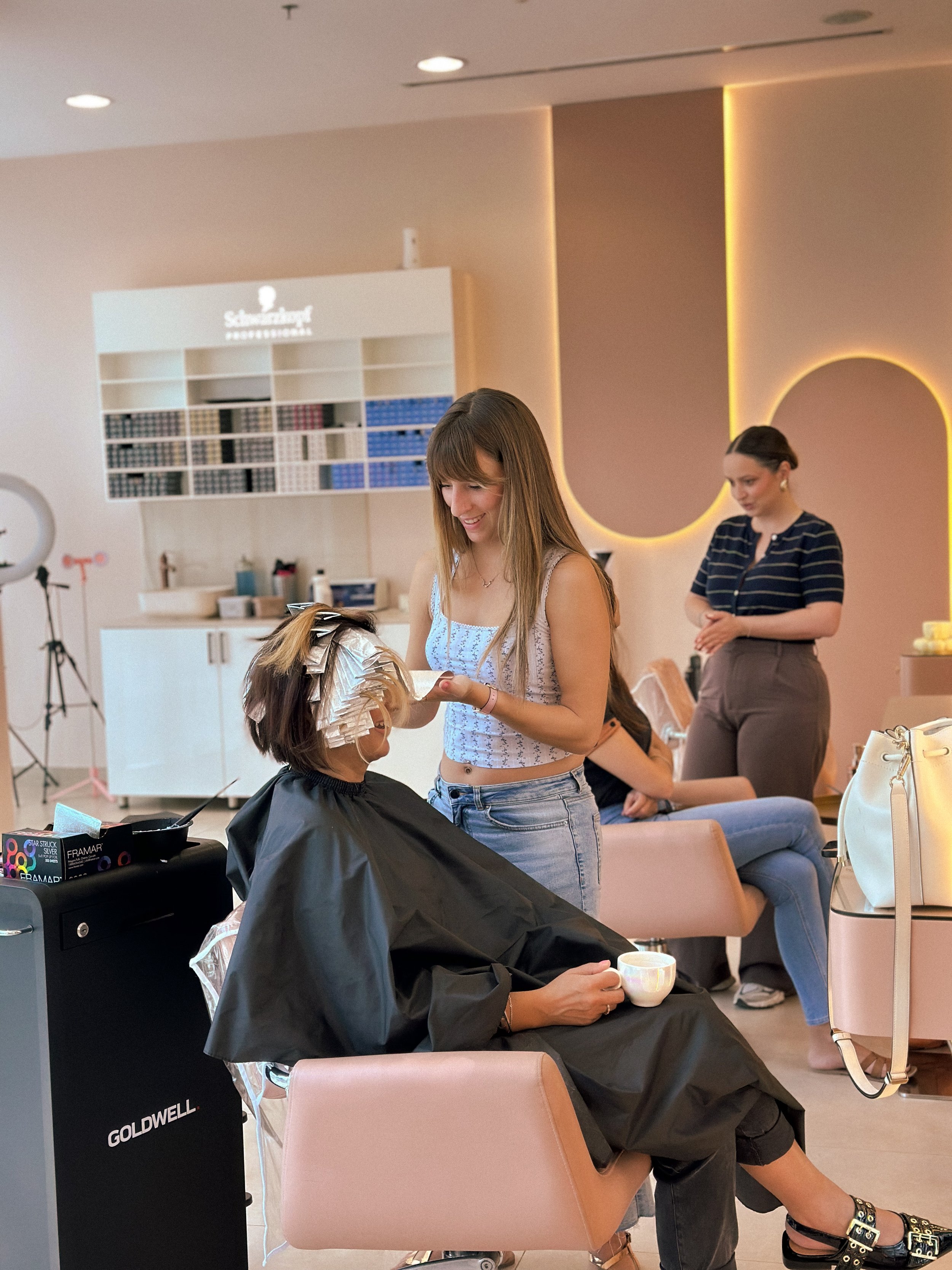 A woman in a salon getting her hair styled with foil highlights, while another woman receives makeup or skincare treatment in the background.