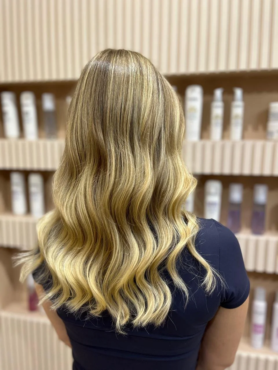 Back view of a woman with long, wavy blonde hair in a salon or store with shelves of hair products.