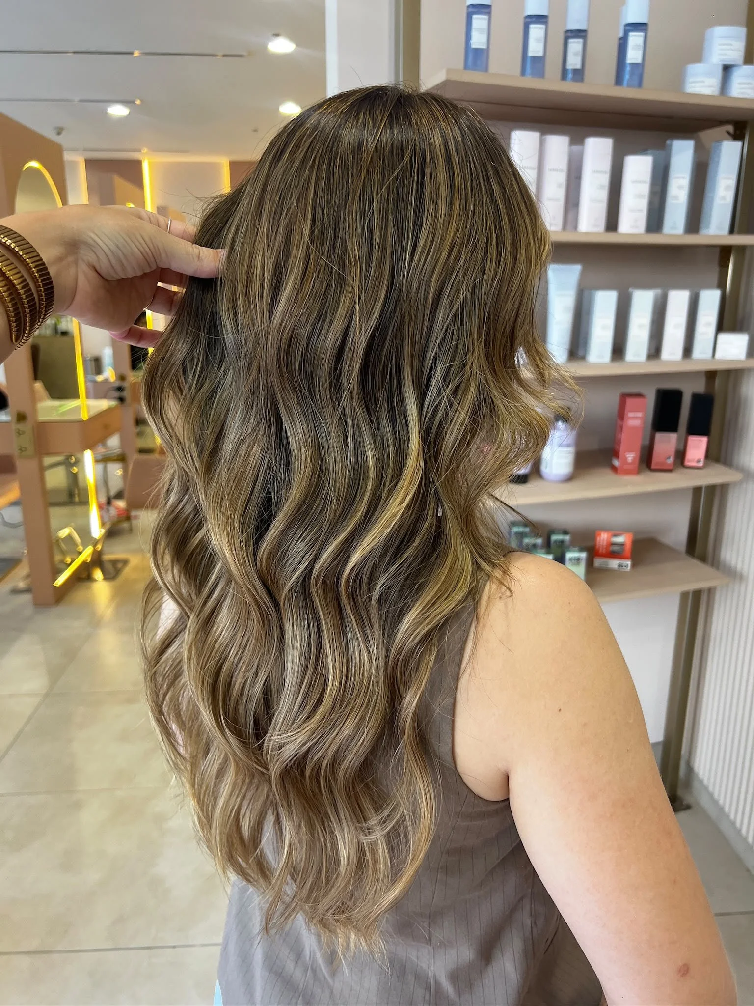 Back view of a woman with long, wavy hair styled in loose curls with blonde highlights, in a salon setting with shelves of hair products in the background.