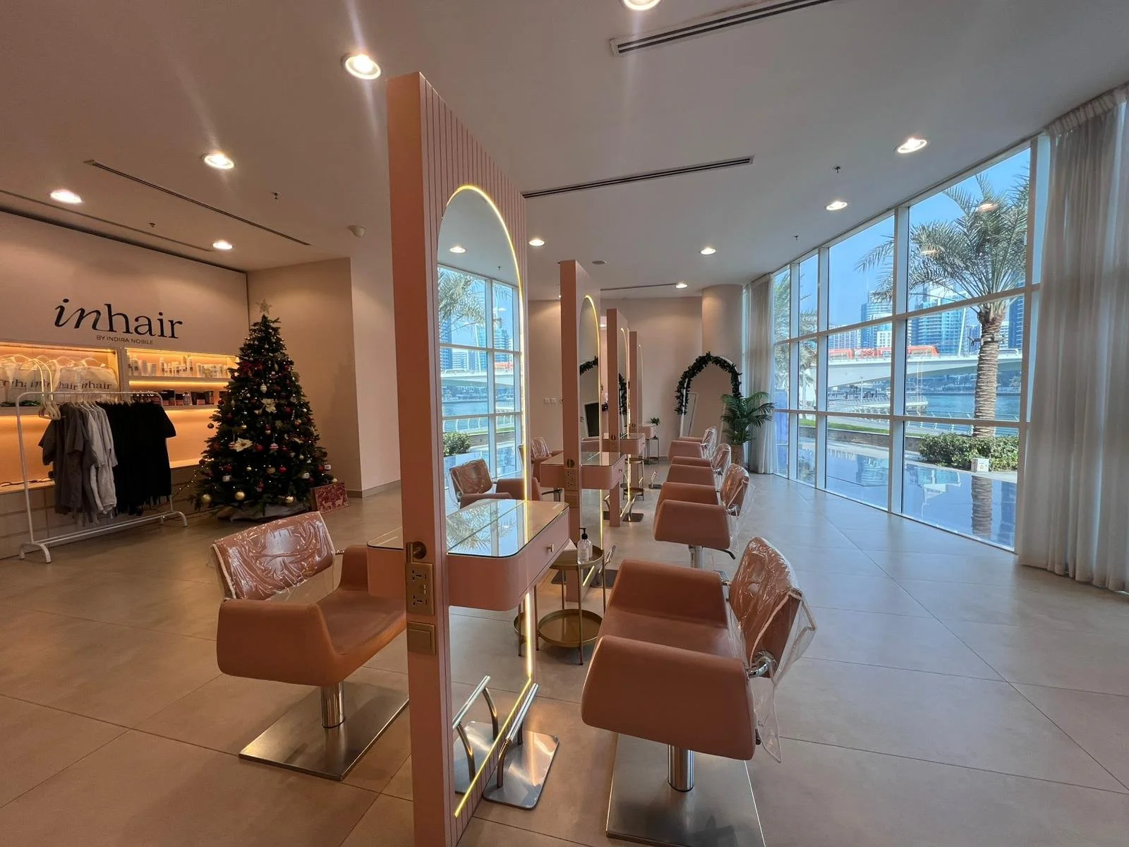 Interior of a hair salon with pink chairs and large windows showing palm trees outside. Christmas tree and holiday decorations inside.