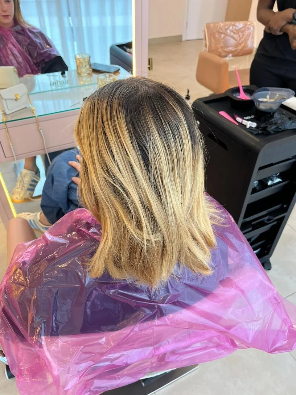 Overhead view of a woman with shoulder-length blonde hair sitting in a salon chair, covered with a pink plastic cape, with hair tools and products nearby.