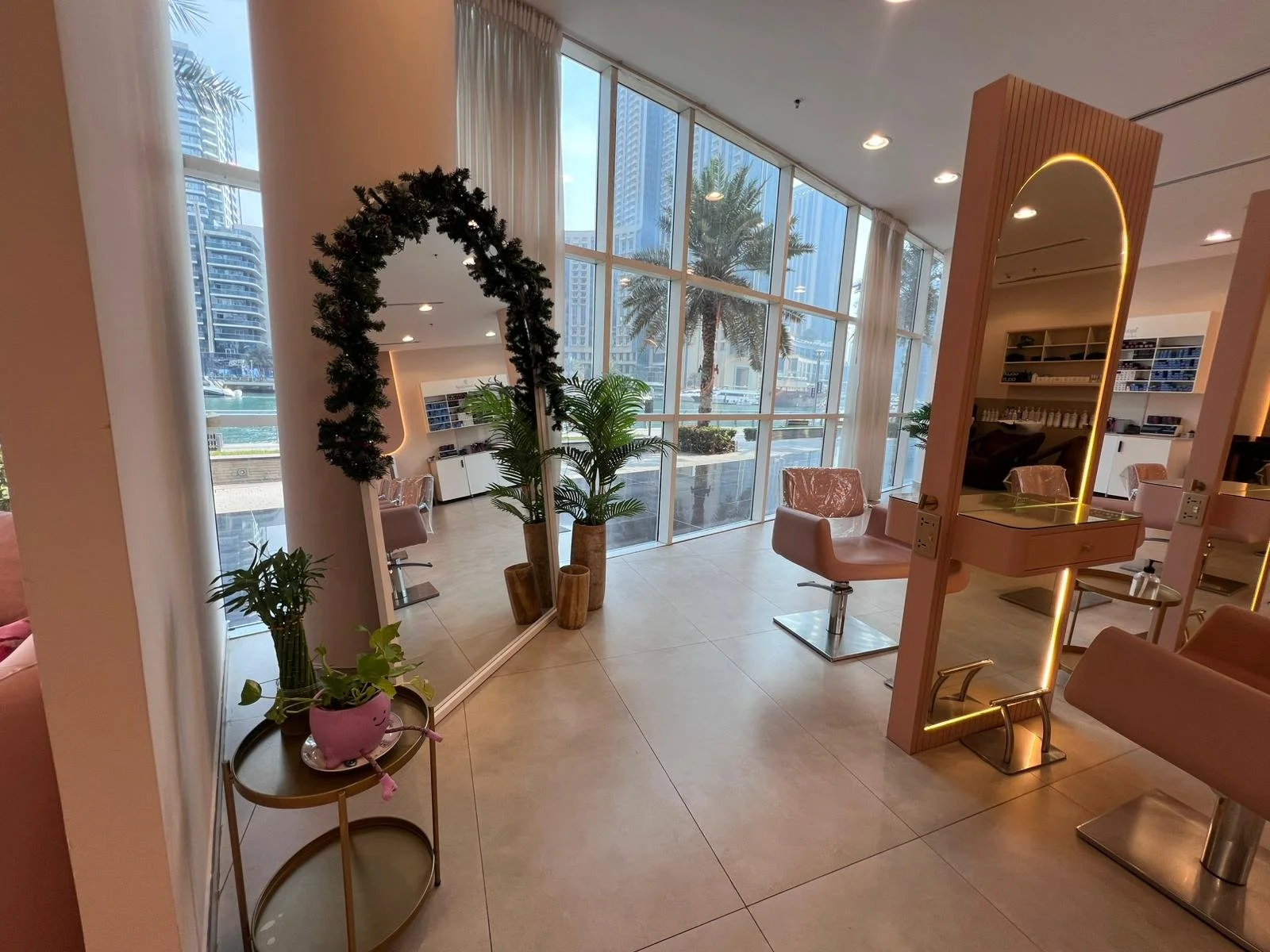 Interior of a modern hair salon with pink chairs, large windows showing city buildings and palm trees outside, potted plants, and a mirror with warm lighting.