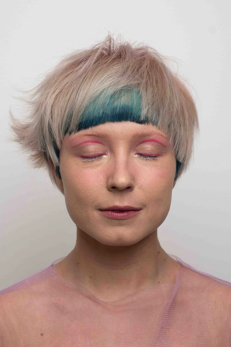 A woman with short, tousled pastel pink hair featuring a strip of dark teal and blonde highlights. She has pink, artistic line makeup above her eyelids, closed eyes, and a serene expression. She wears a light pink, sheer top.