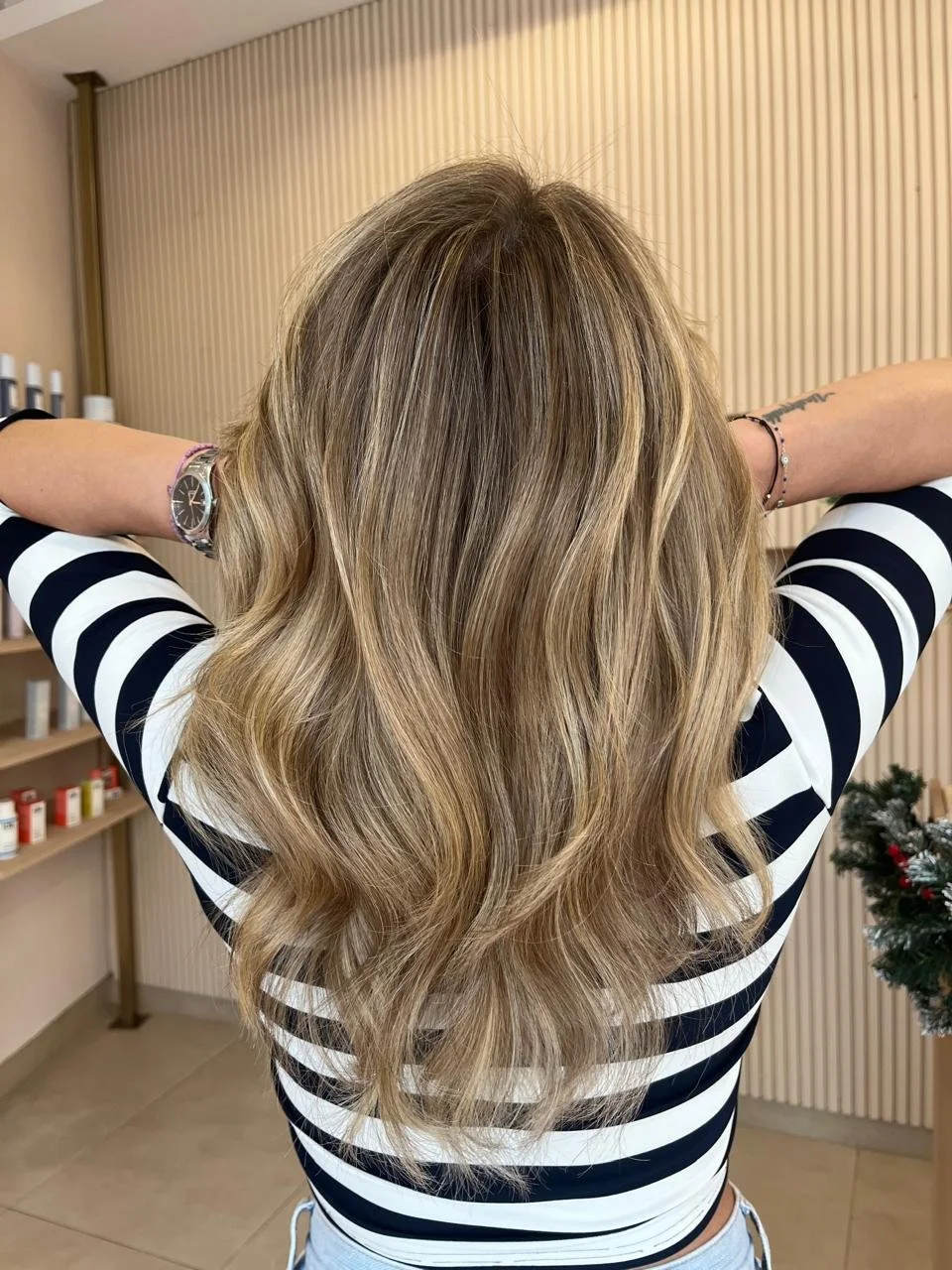 Back view of woman with long, wavy, blonde hair, wearing a black and white striped shirt, in a salon or similar setting.