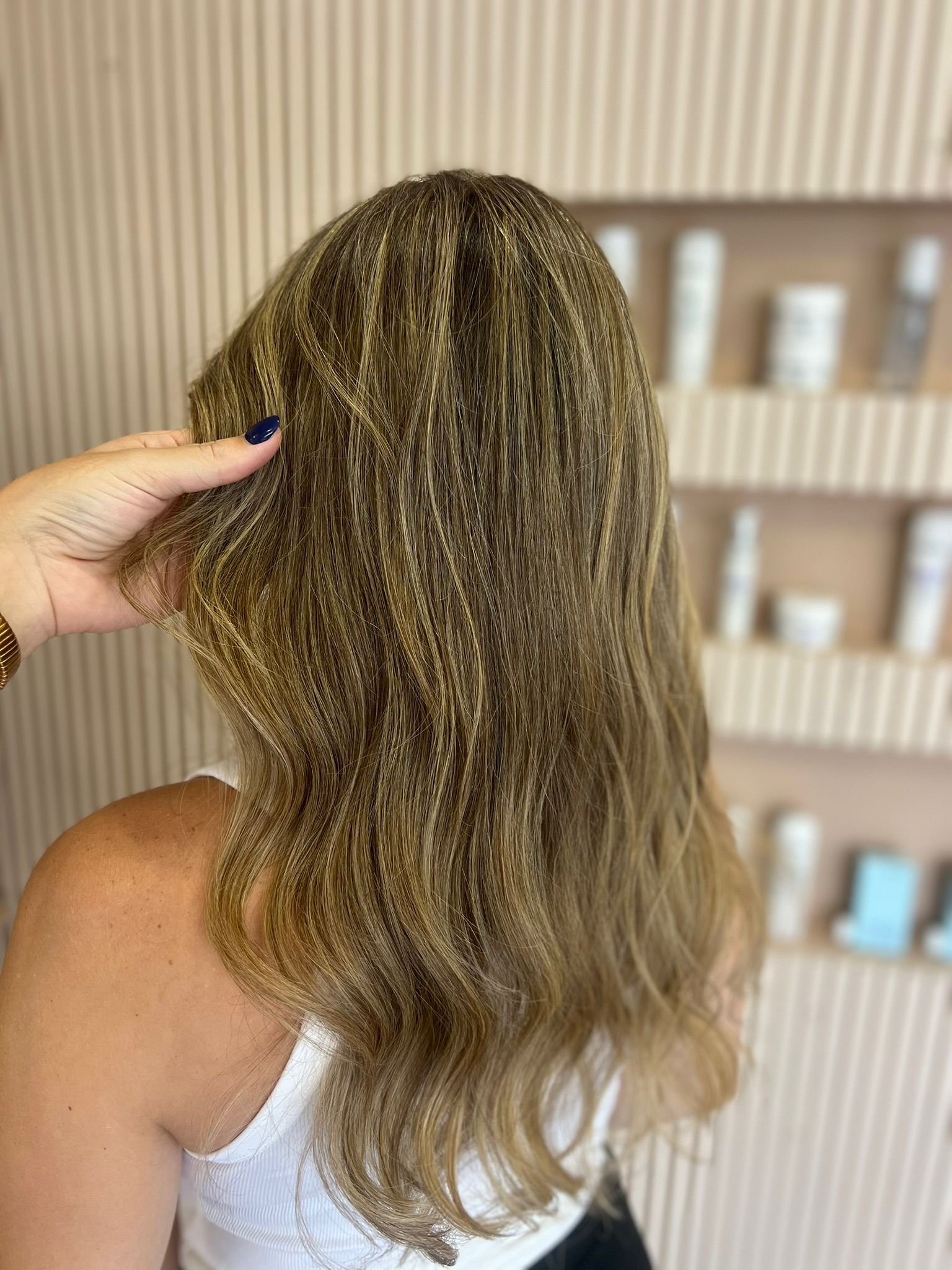 A person with long, wavy blonde hair wearing a white sleeveless top, with a hand touching the hair on the side of the head, standing in front of a beige, textured wall with shelves holding skincare or haircare products.