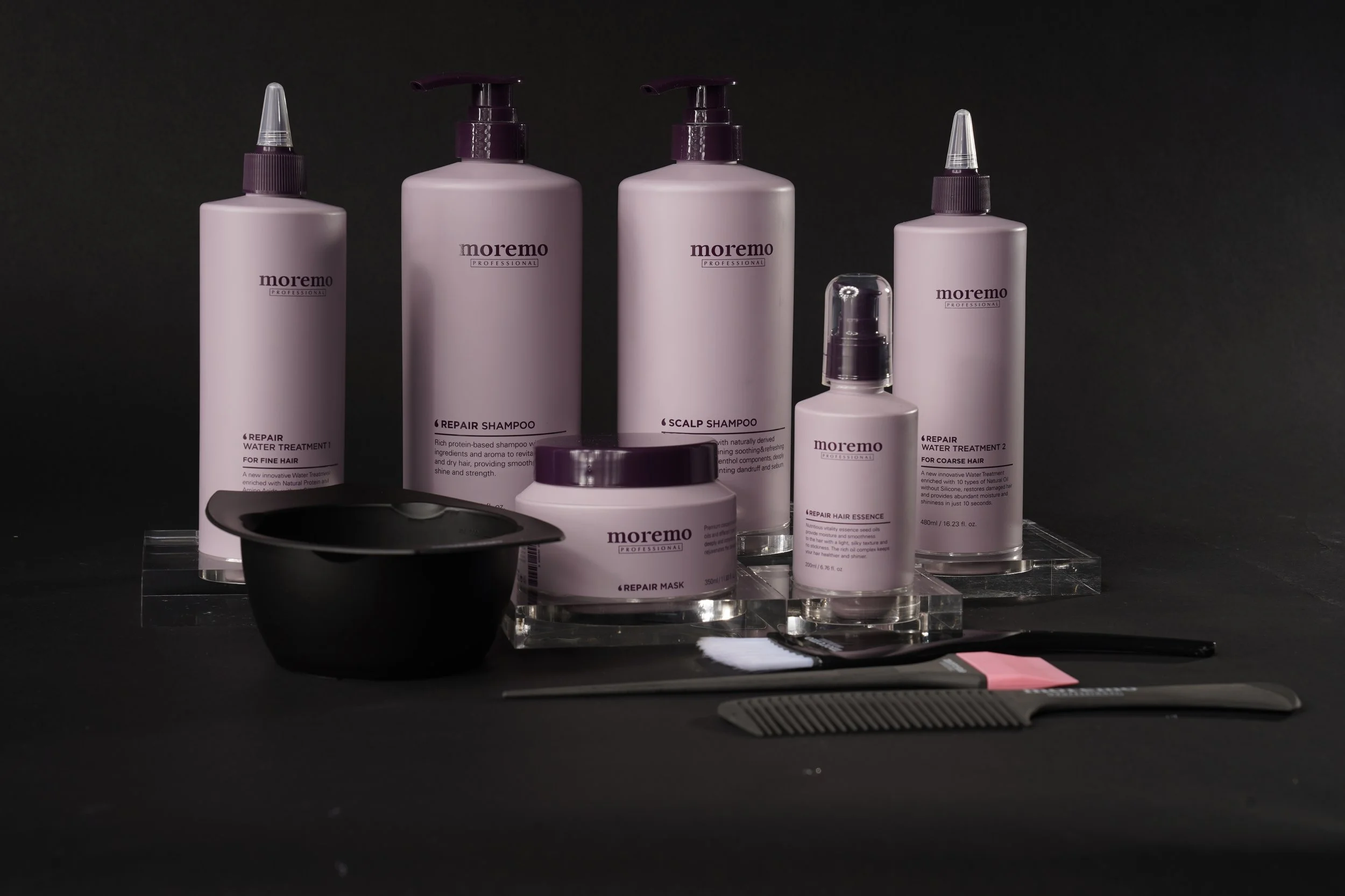 Collection of pink hair care products including shampoo, conditioner, mask, and styling sprays, along with hairbrushes and a bowl, on a black background.