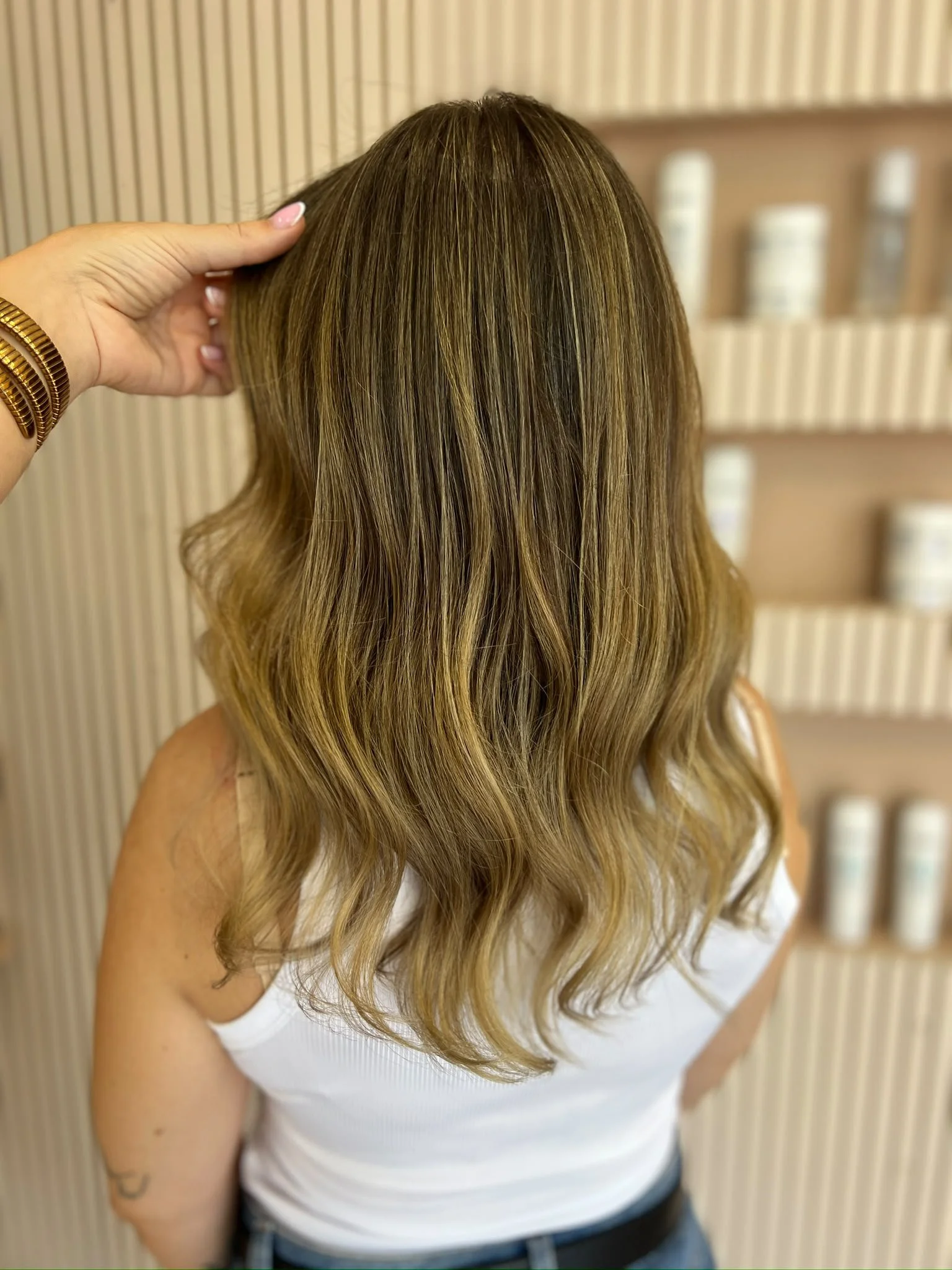 Back view of a woman with long, wavy, light brown hair wearing a white tank top, standing in front of a wooden wall with shelves of hair care products.