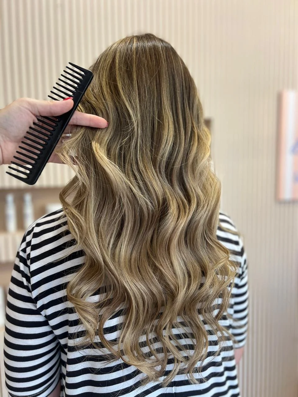 Person with wavy blonde hair sitting in a salon, with a hand holding a wide-tooth comb and pointing to the hair.