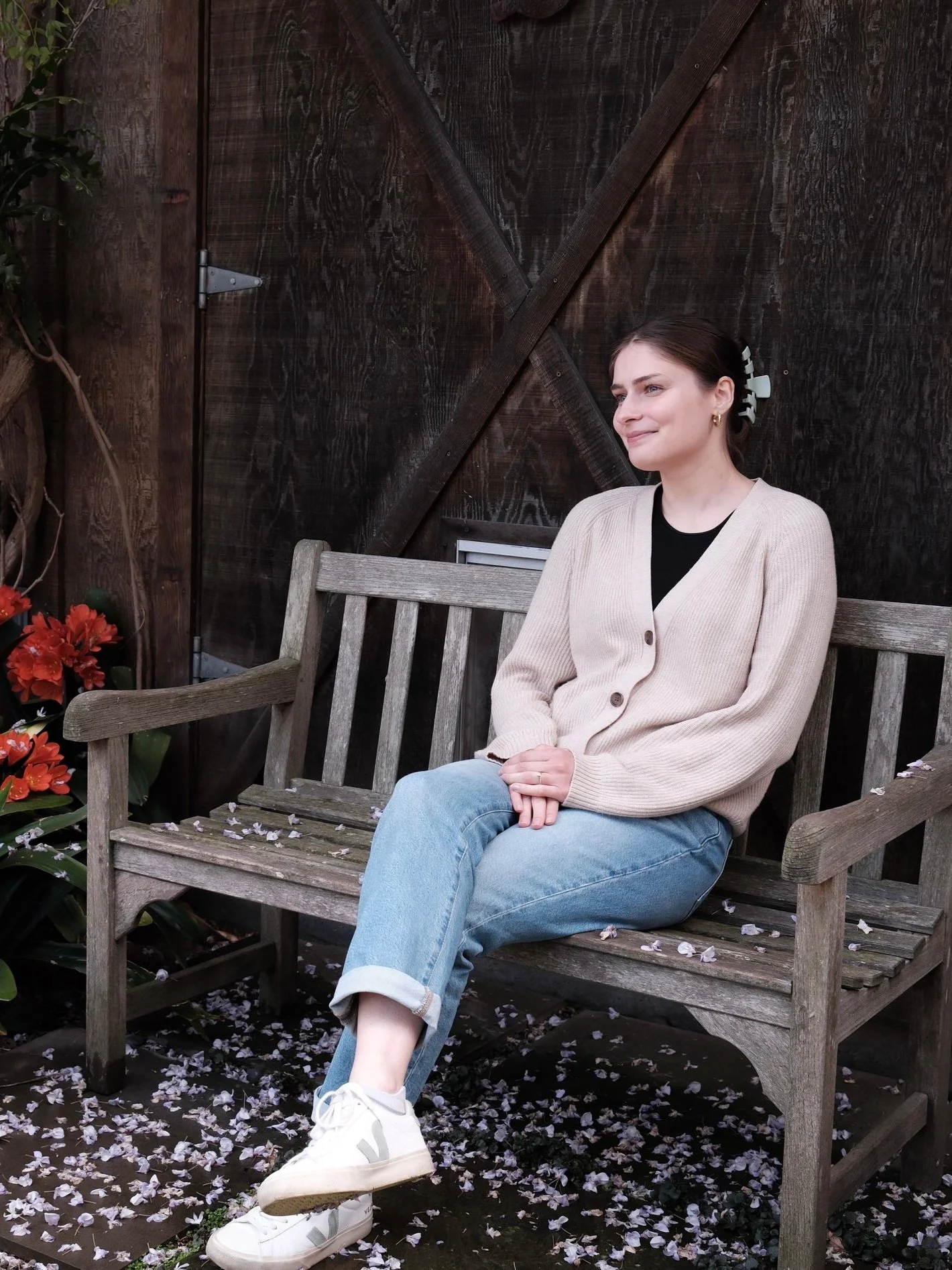 A woman sitting on a wooden garden bench with fallen flower petals around her, dressed in a beige cardigan, black shirt, and light blue jeans, with a hair clip in her hair, smiling softly.