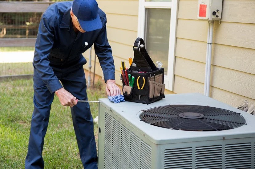 A technician wearing blue uniform and cap cleaning an outdoor air conditioning unit with a brush and cloth.