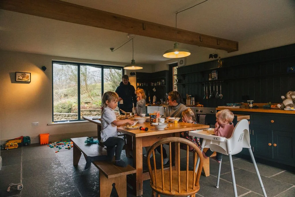 A group of children and an adult sitting around a wooden dining table in a kitchen, with toys on the floor and a large window showing an outdoor scene with trees.