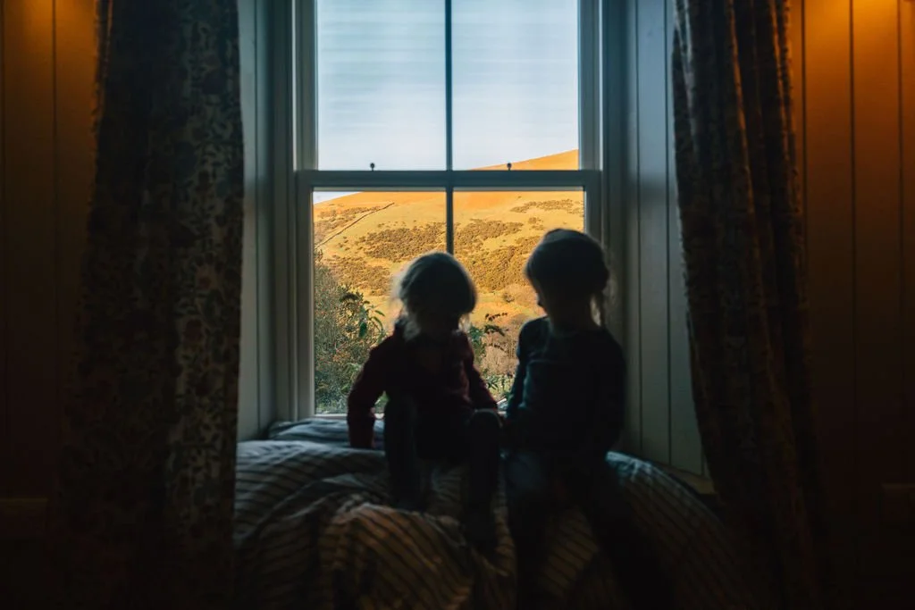 Two children sitting on a window seat, silhouetted against a scenic outdoor view of rolling green hills and a partly cloudy sky, framed by curtains.