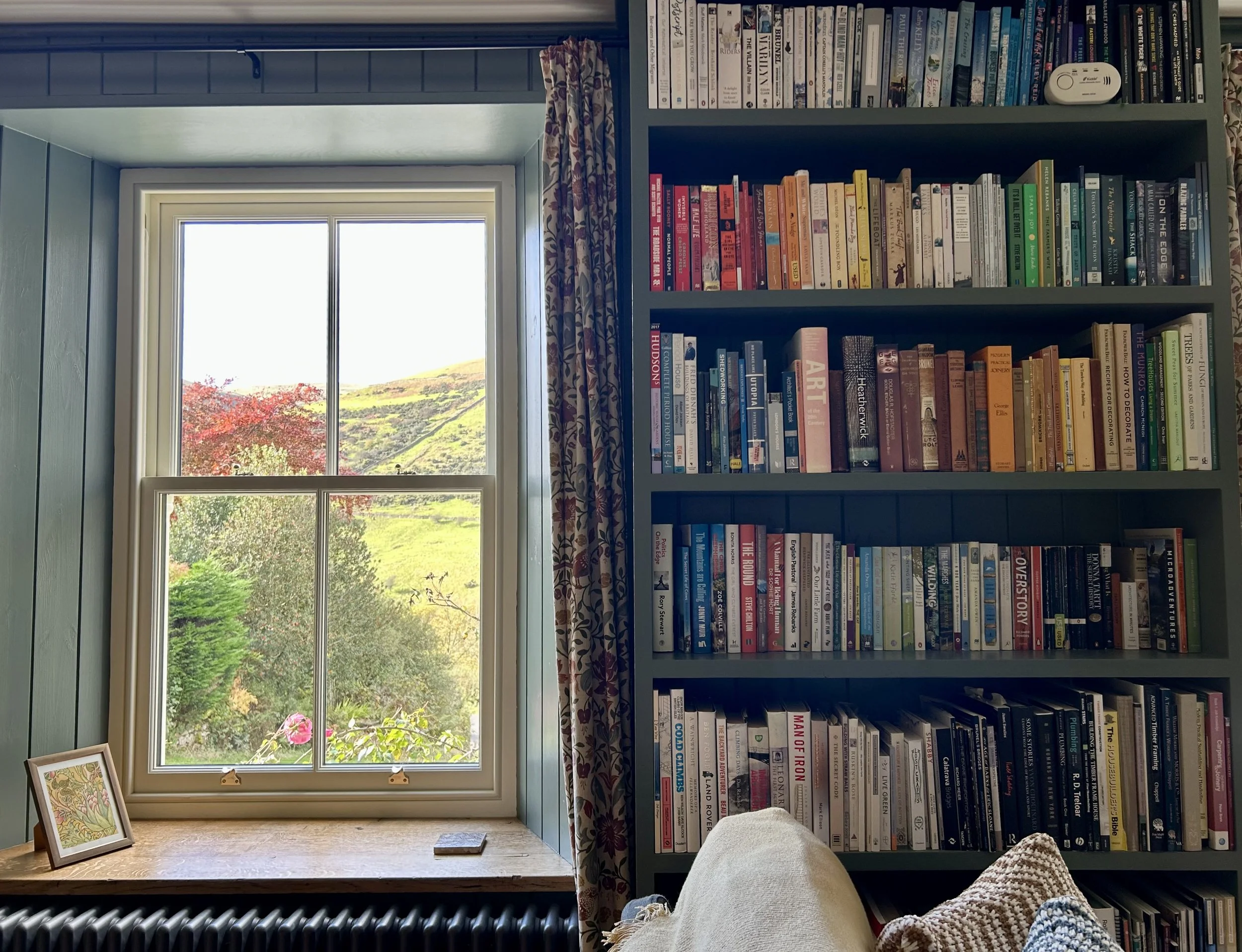 A cozy room with a window overlooking a scenic landscape of trees and hills. Inside, a wooden desk underneath the window has a small framed picture and a book. To the right, a tall bookshelf filled with colorful books is against the wall, and part of a person's leg with a sweater is visible in the bottom right corner.