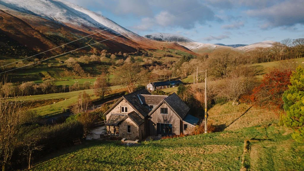 A rustic house in a green hilly area with mountains in the background, some snow on the peaks, and trees with autumn foliage.