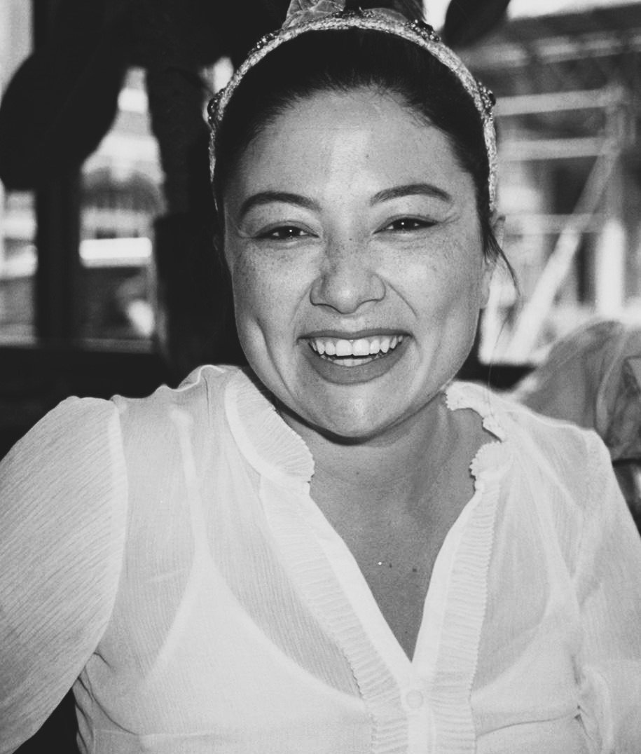 Black and white photo of a woman smiling with dimples, wearing a headband and a light-colored blouse, in an outdoor or semi-outdoor setting.