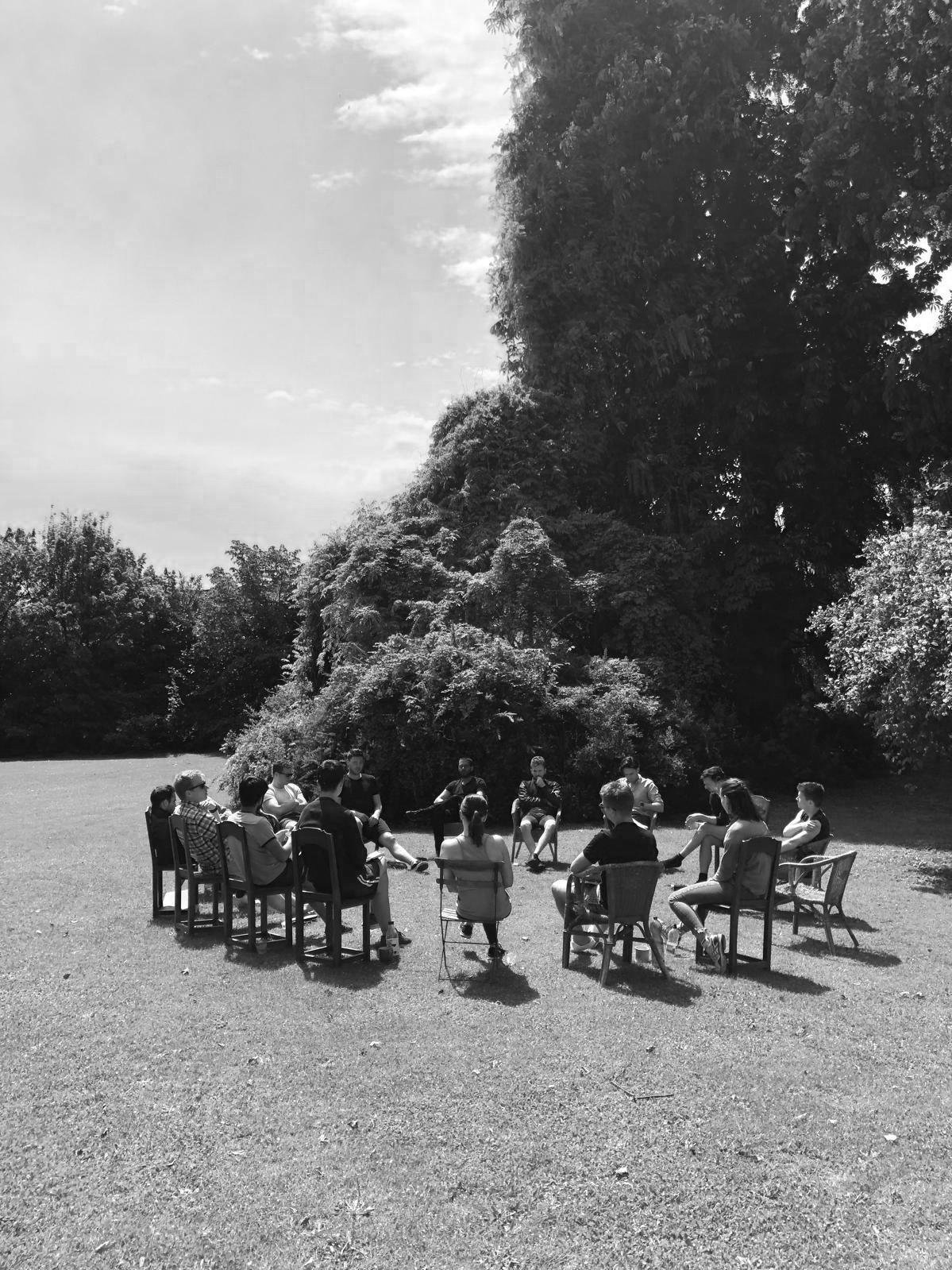 Group of people sitting in a circle outdoors on grass, facing each other, with trees in the background.