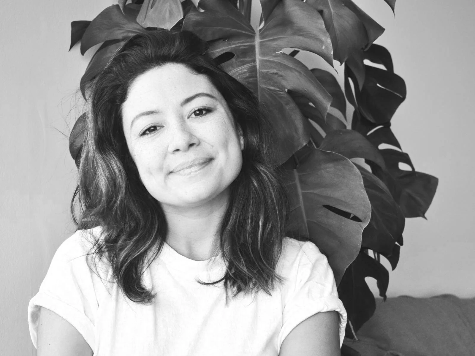 Black and white photo of a woman with wavy hair smiling, sitting in front of a large leafy plant, wearing a light-colored shirt.