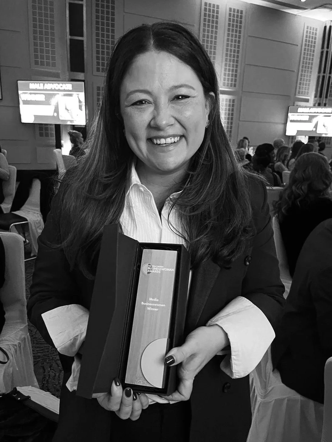A woman smiling and holding an award plaque at an indoor event with a crowd and screens in the background.